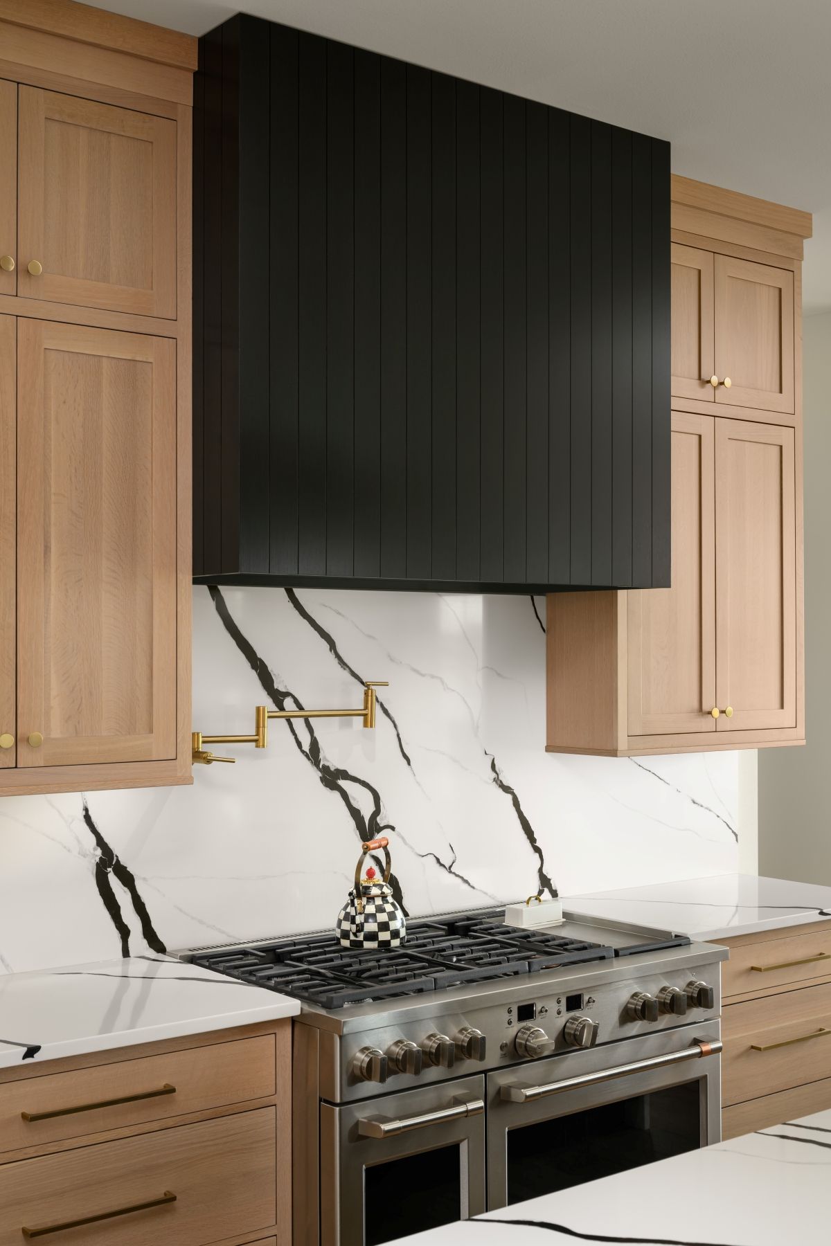 Modern kitchen with black range hood, marble backsplash, stainless stove, and light wood cabinets