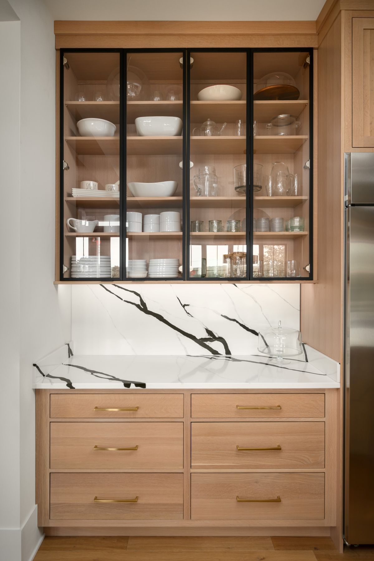 Built-in kitchen cabinet with glass doors, dishes, and wood drawers below a marble backsplash.