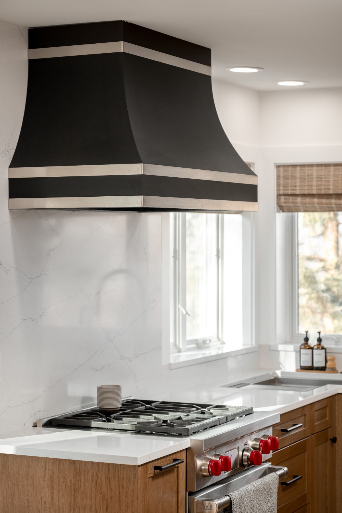 Modern kitchen with black range hood, gas stove, white backsplash, and wood cabinets by a window