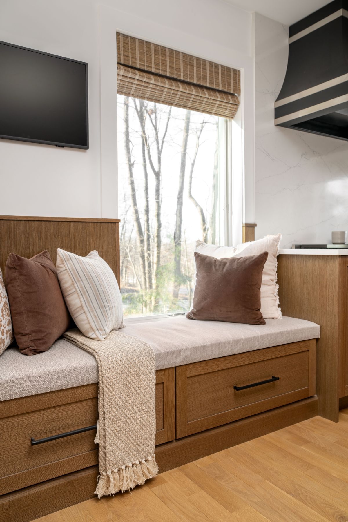 Cozy window bench with brown cushions, white knit throw, wood drawers, and woven shade in a bright room