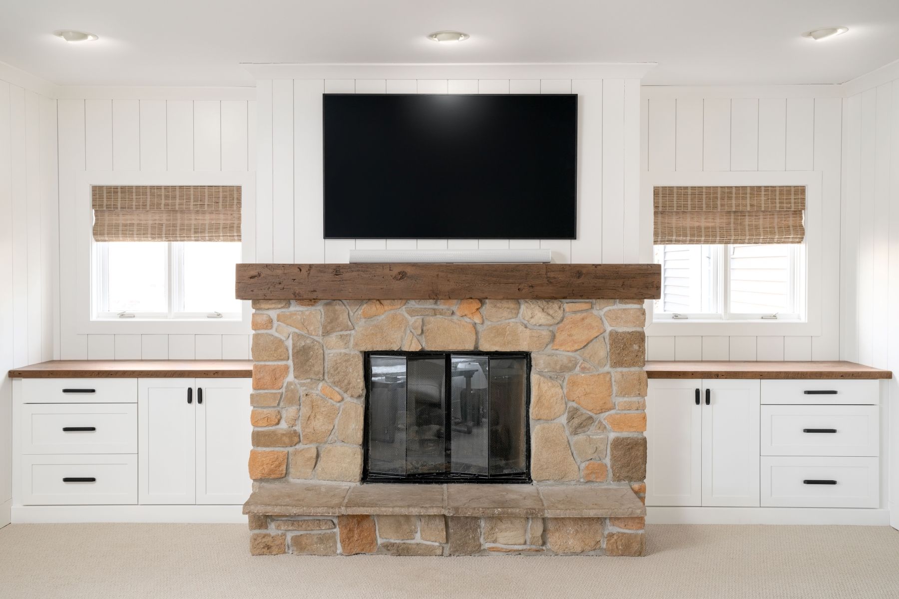 Bright living room with stone fireplace, large TV, white built-ins, and woven window shades