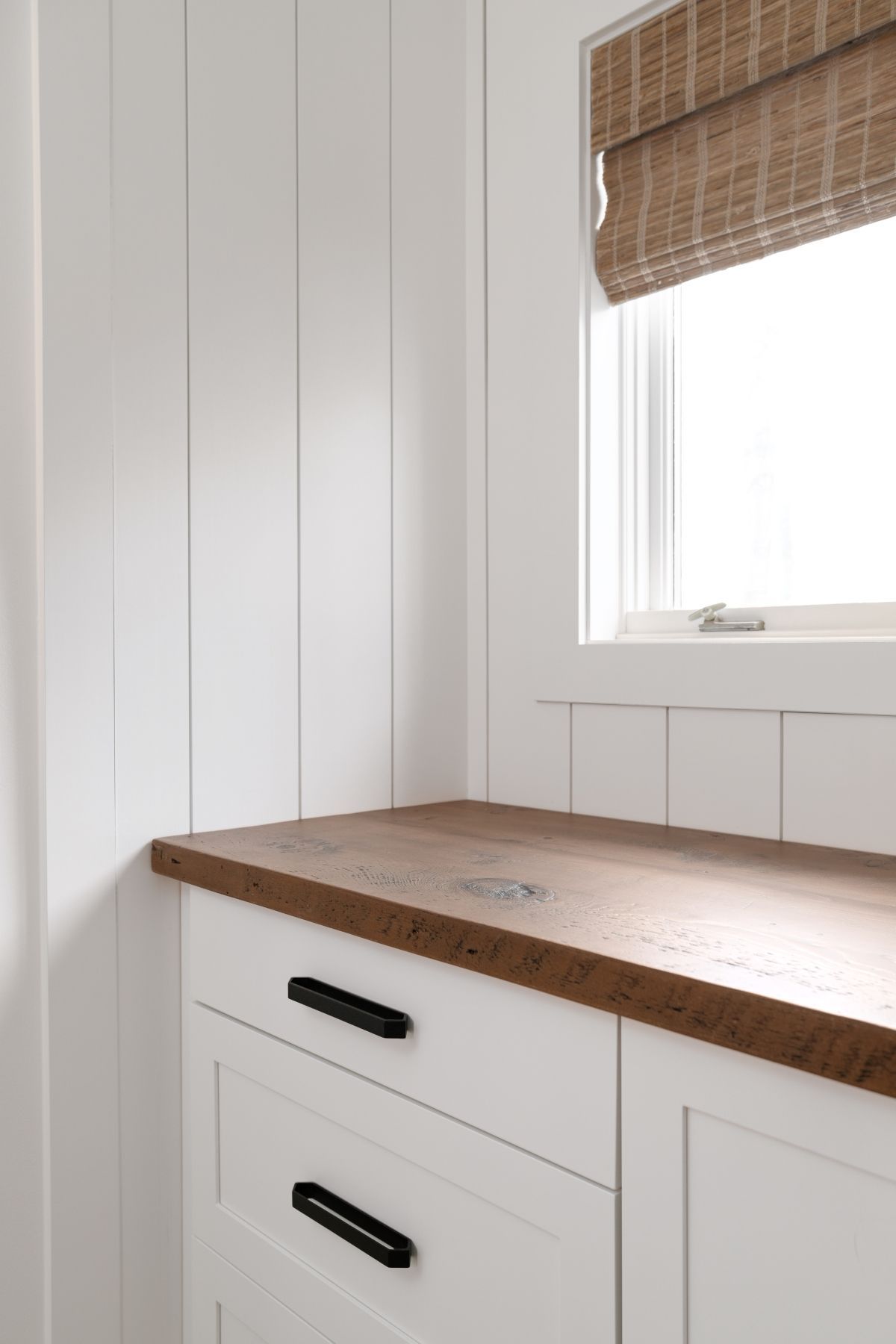 White built-in bench under a window with bamboo blind, next to white paneled walls and a wooden countertop