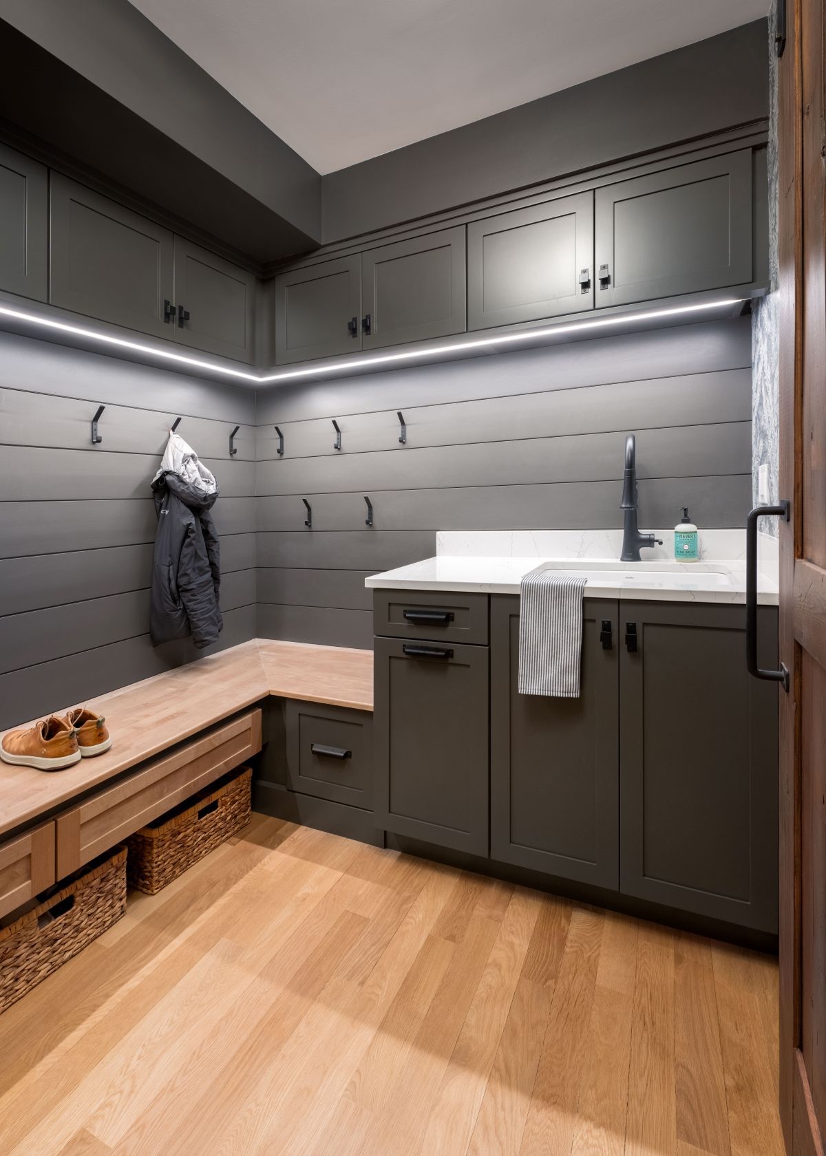 Modern mudroom with gray cabinets, built-in bench, hooks, and white countertop sink under under-cabinet lighting