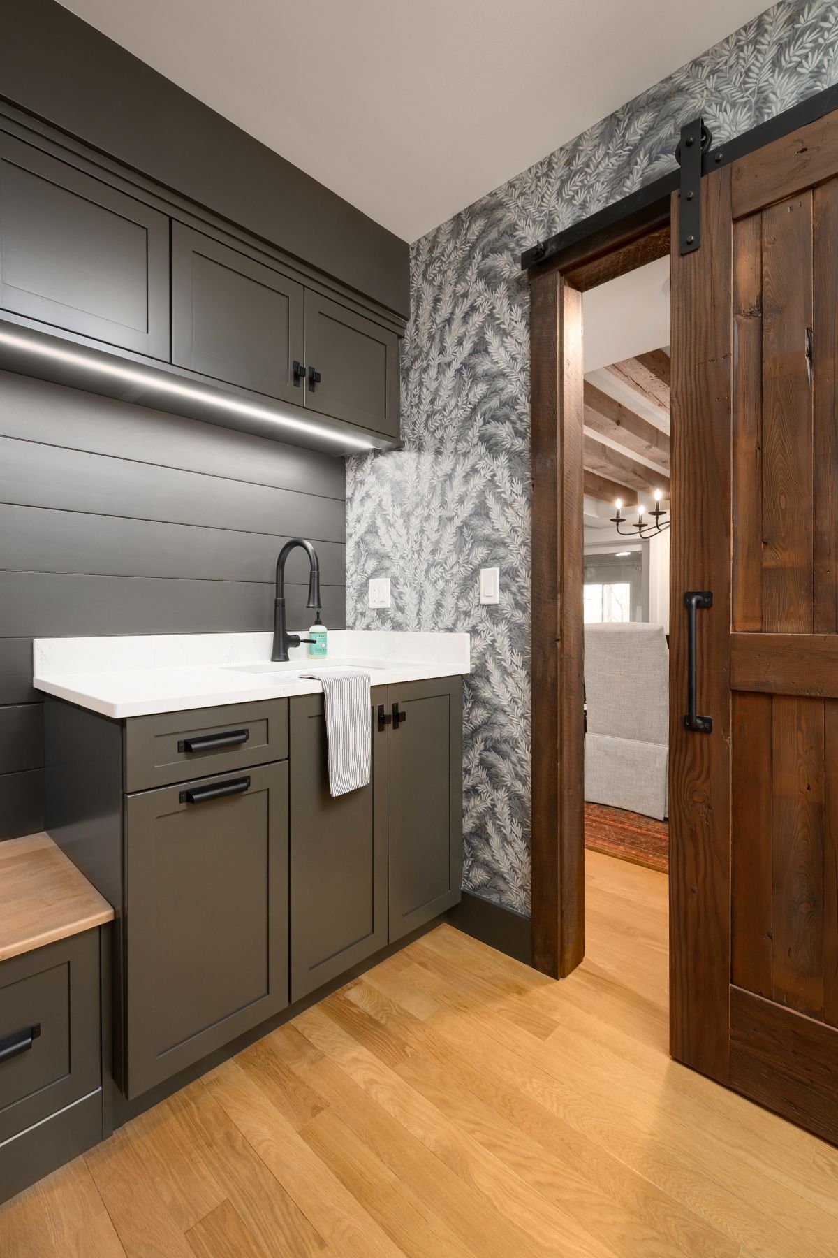 Modern kitchen sink area with dark cabinets, white countertop, wood floor, and open wooden door to hallway
