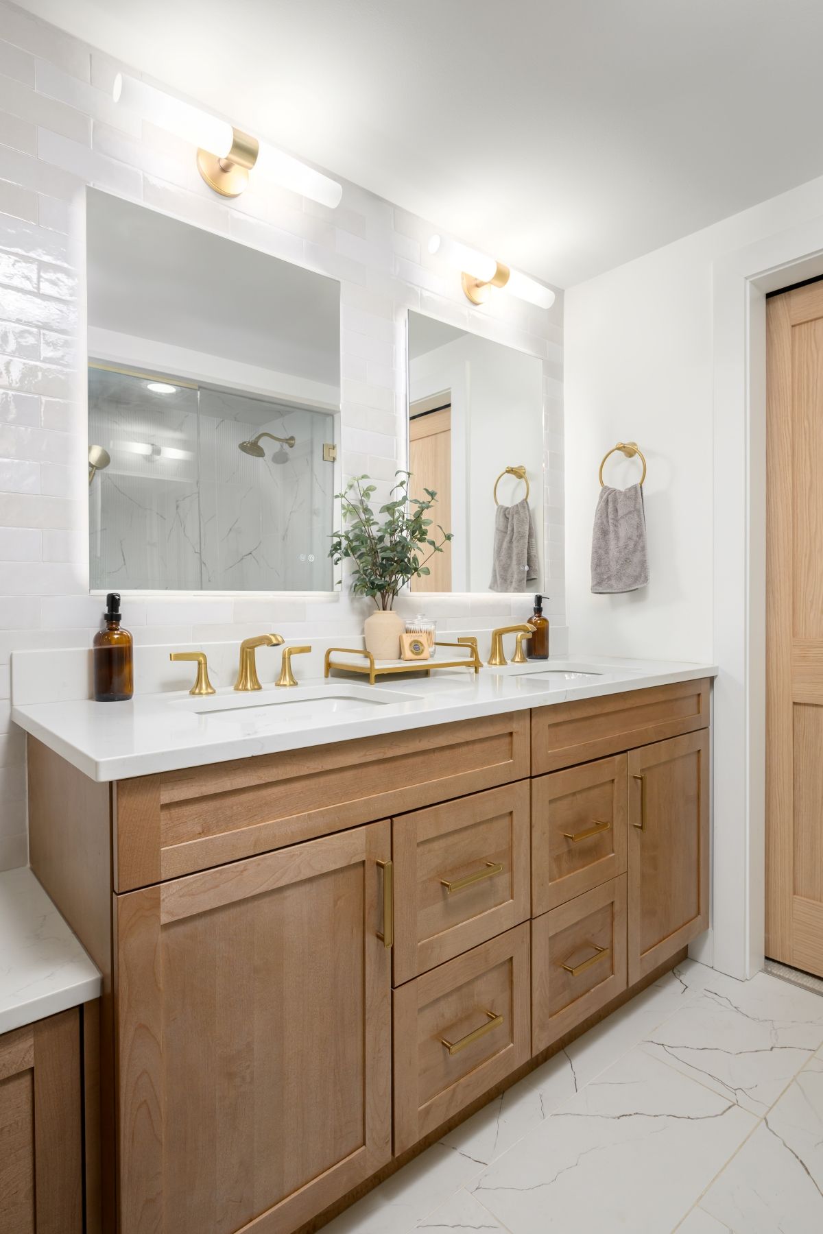 Modern bathroom vanity with wood cabinets, double mirrors, gold fixtures, and white tile floor