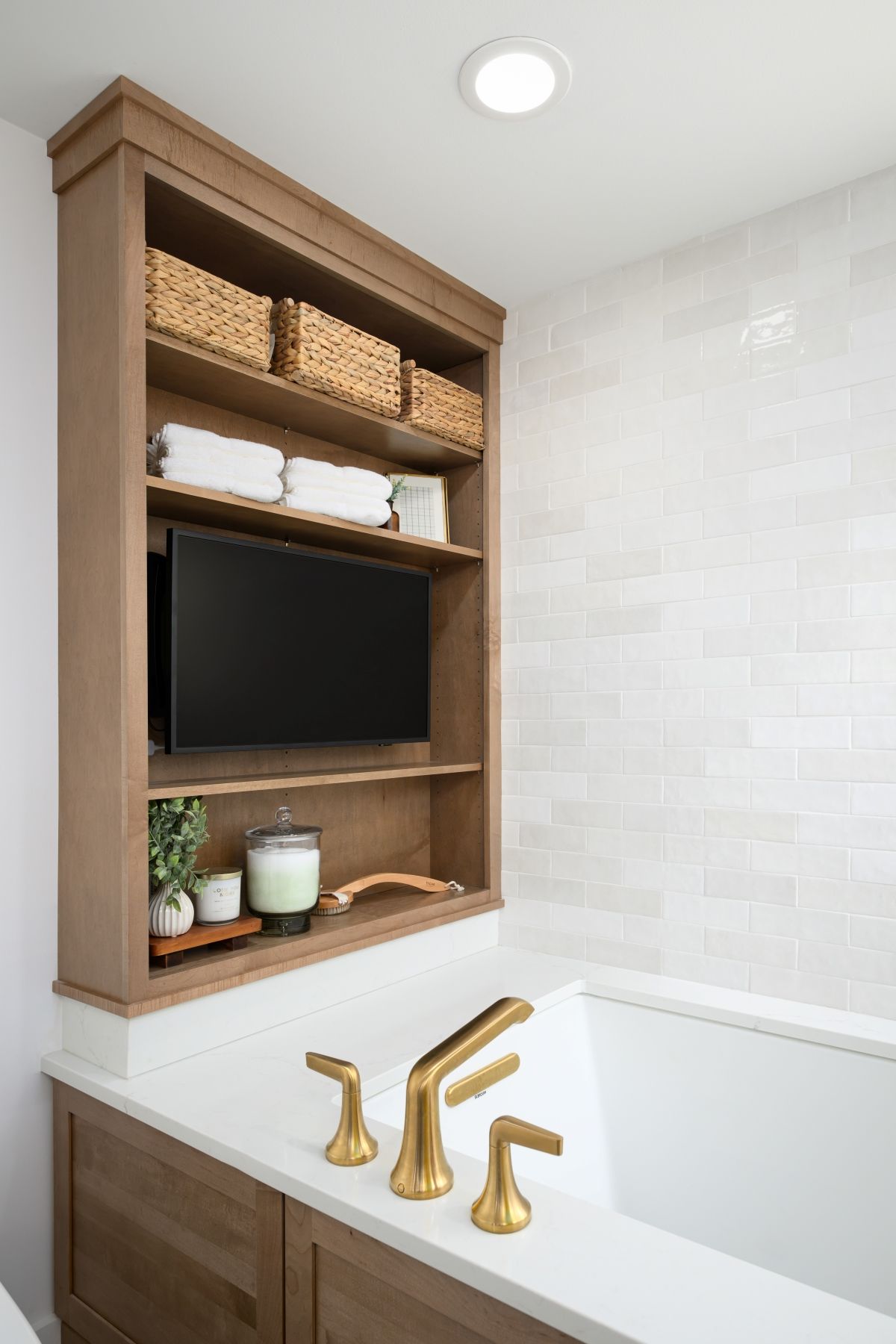 Bathroom vanity with gold faucet, built-in shelving, baskets, towels, and wall-mounted TV over the tub