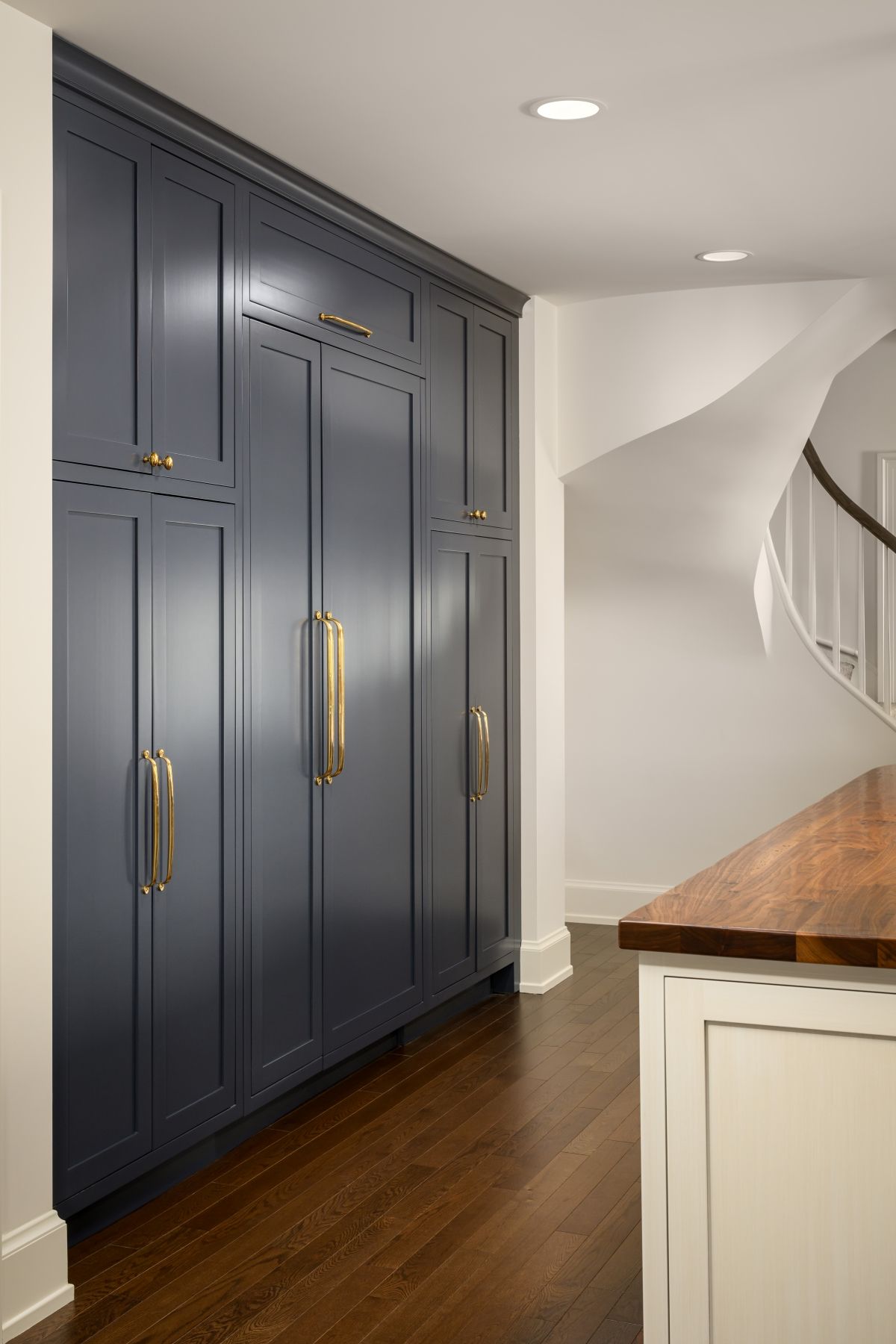 Dark blue built-in cabinets with gold handles in a bright hallway beside a wood countertop and staircase.