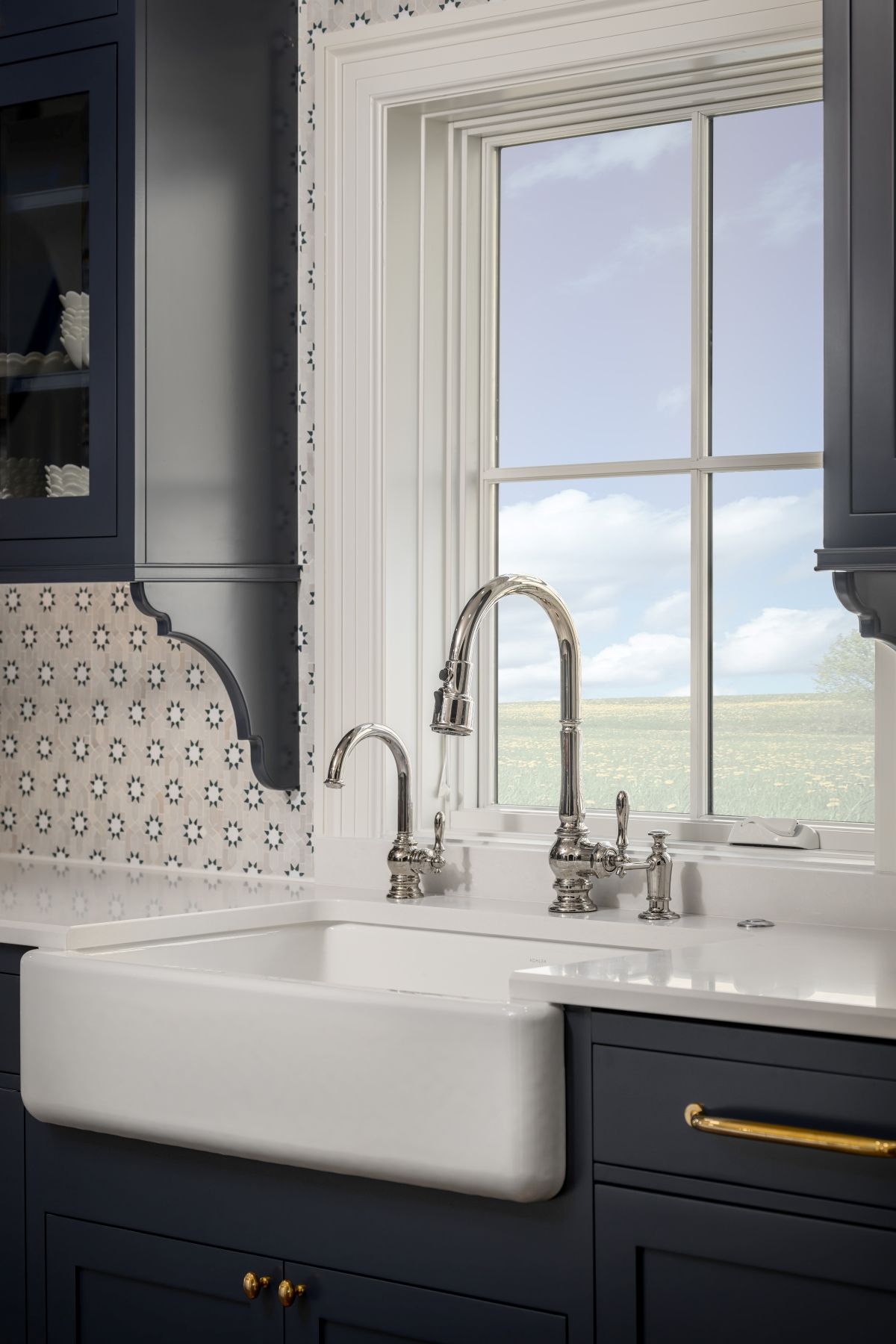 Modern kitchen sink with white apron-front basin, brass faucet, and window view of cloudy sky and field