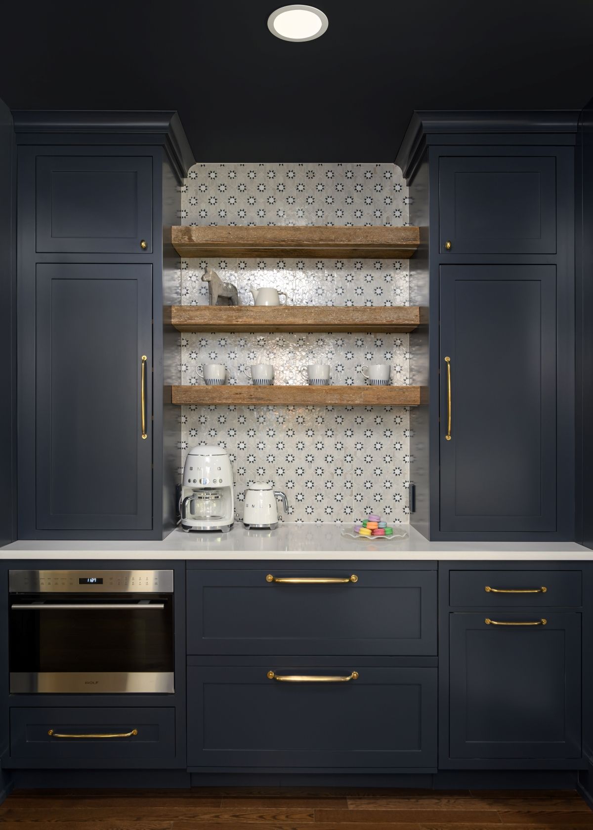 Dark navy kitchen cabinets with brass handles, open shelves, white patterned backsplash, and built-in oven