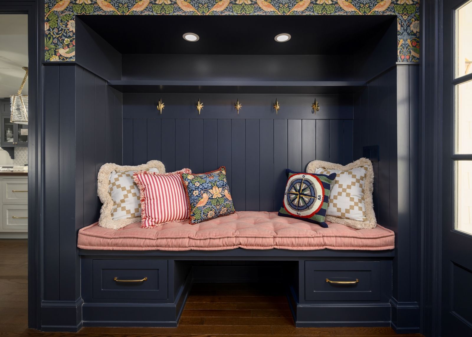 Navy built-in bench nook with pink cushion, patterned pillows, brass hooks, and small overhead light