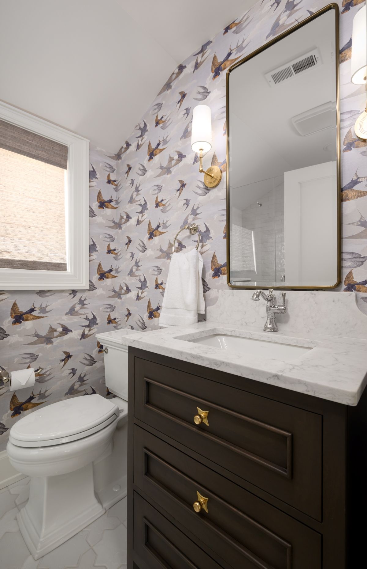 Compact bathroom with bird wallpaper, dark vanity, white countertop, mirror, toilet, and window shade.