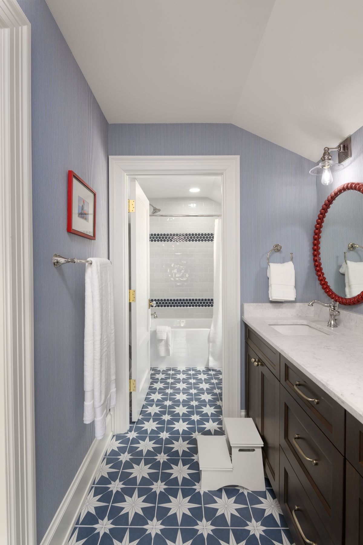 Narrow bathroom with blue striped wallpaper, star-pattern tile floor, dark vanity, and red-framed mirror.