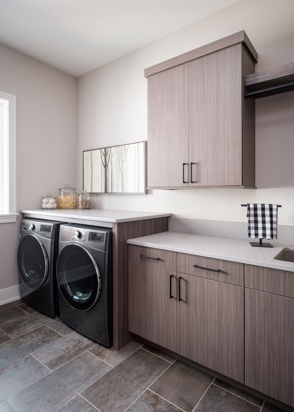 Modern laundry room with front-load washer and dryer, wood cabinets, gray tile floor, and countertop.