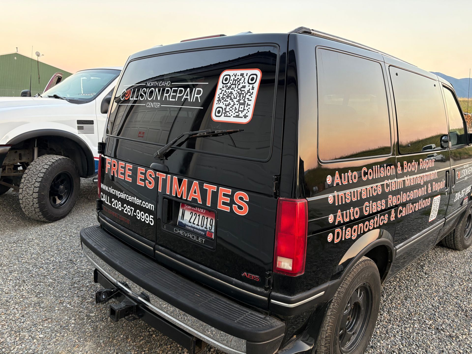Black van with service signage and QR code, parked next to a white truck outdoors.