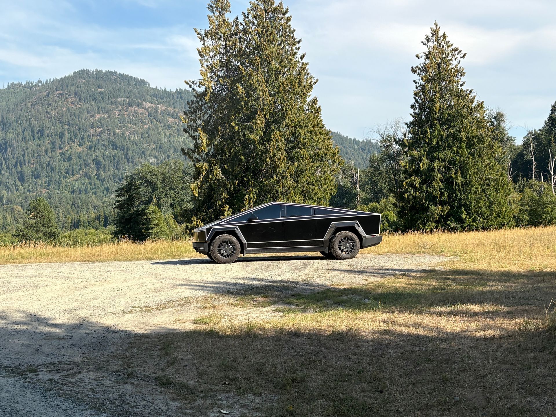 Black Tesla Cybertruck parked on gravel with a mountain backdrop and trees.