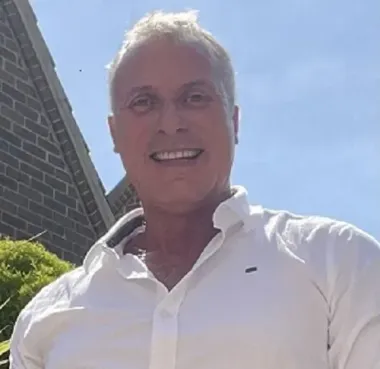 A man in a white shirt is smiling in front of a brick building.