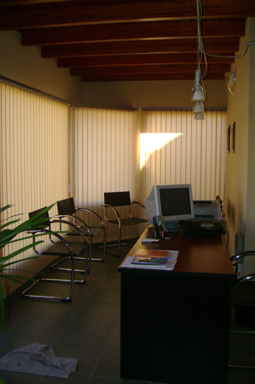 Pequeña oficina con escritorios, sillas, persianas y una computadora cerca de una ventana soleada.