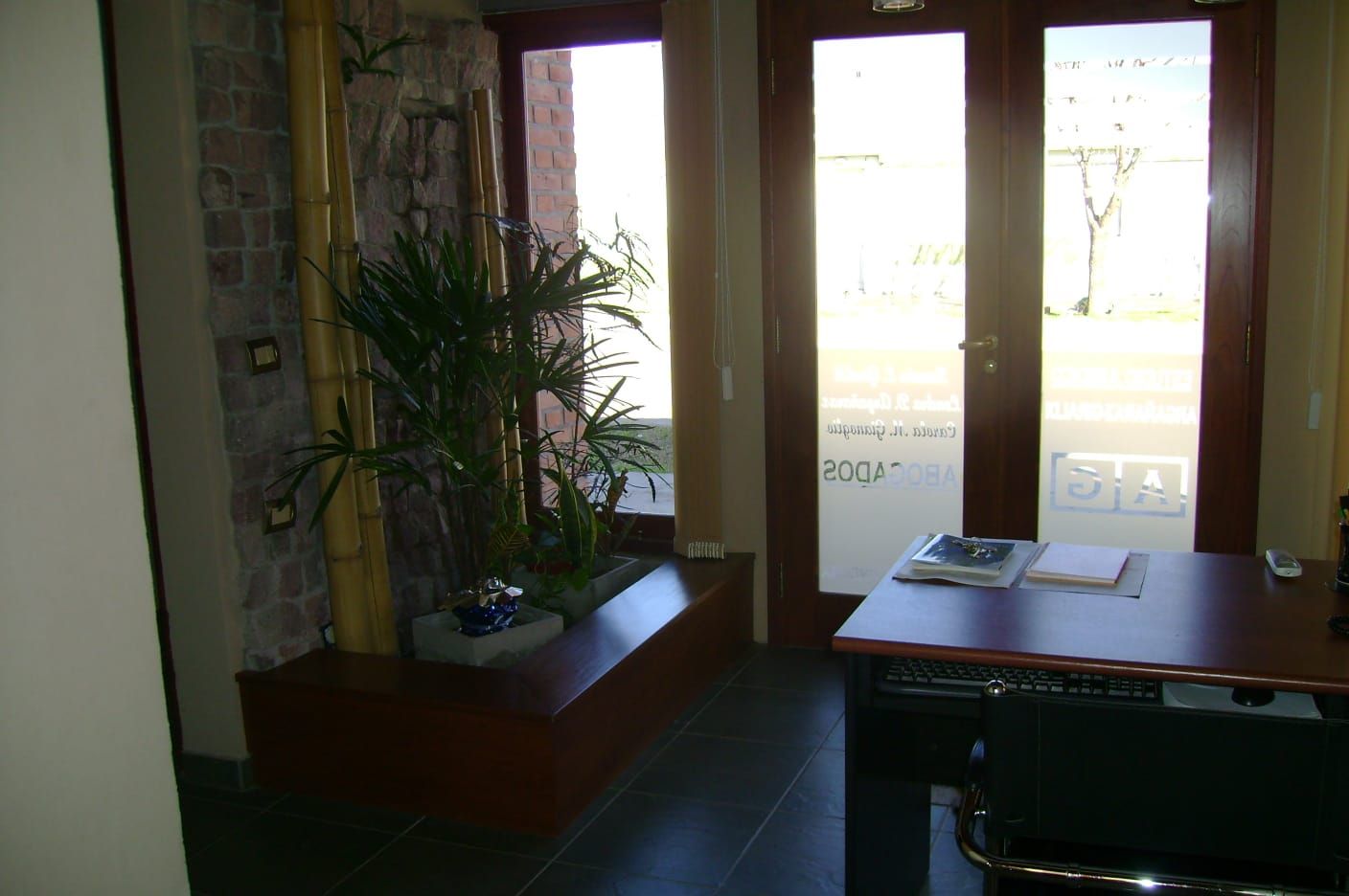 Interior con poca luz, con una planta en maceta, una mesa y puertas de cristal brillante que dan al exterior.