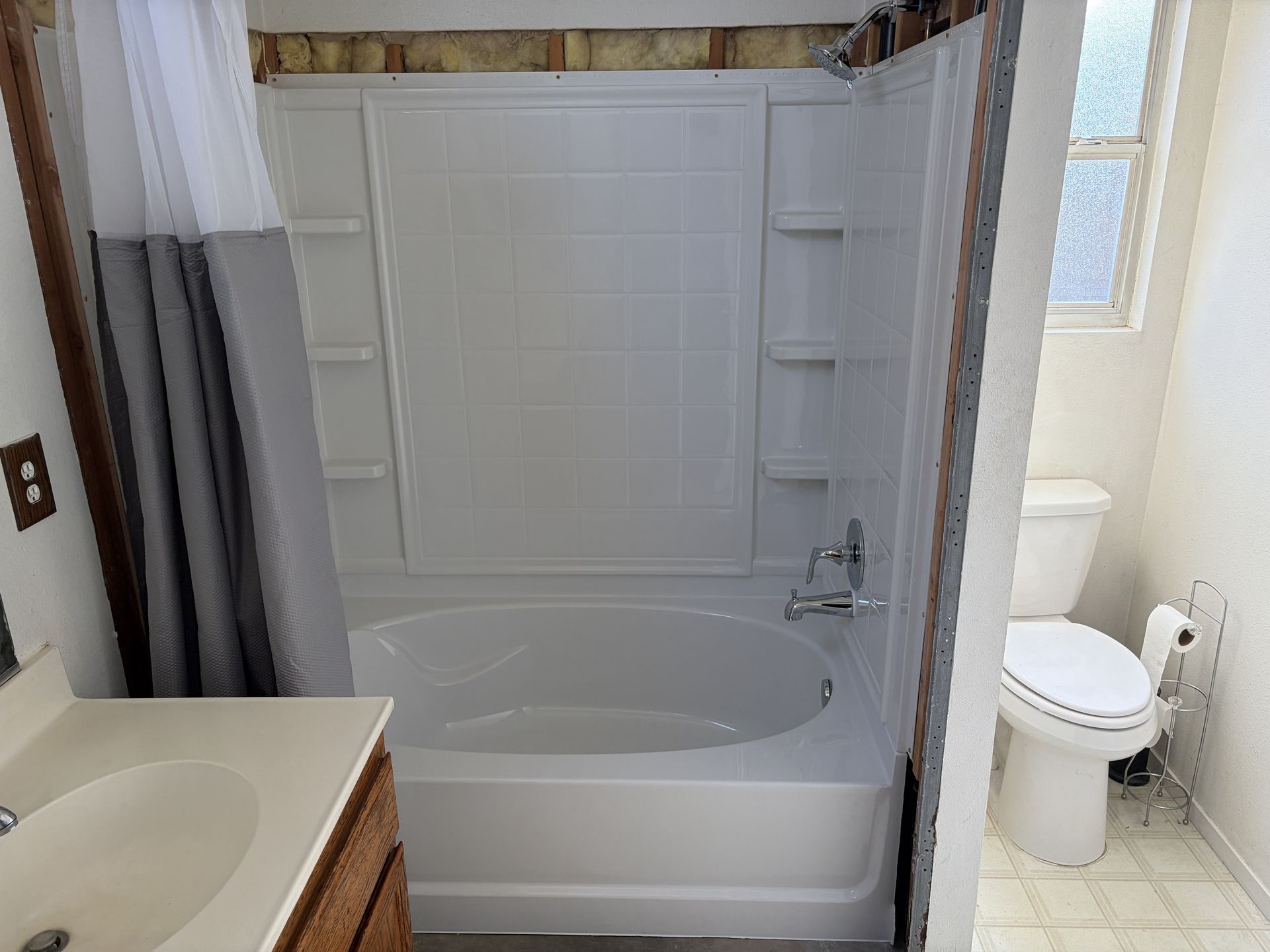 A white bathtub and shower surround with an unfinished, exposed wall above it, next to a toilet in a small bathroom.
