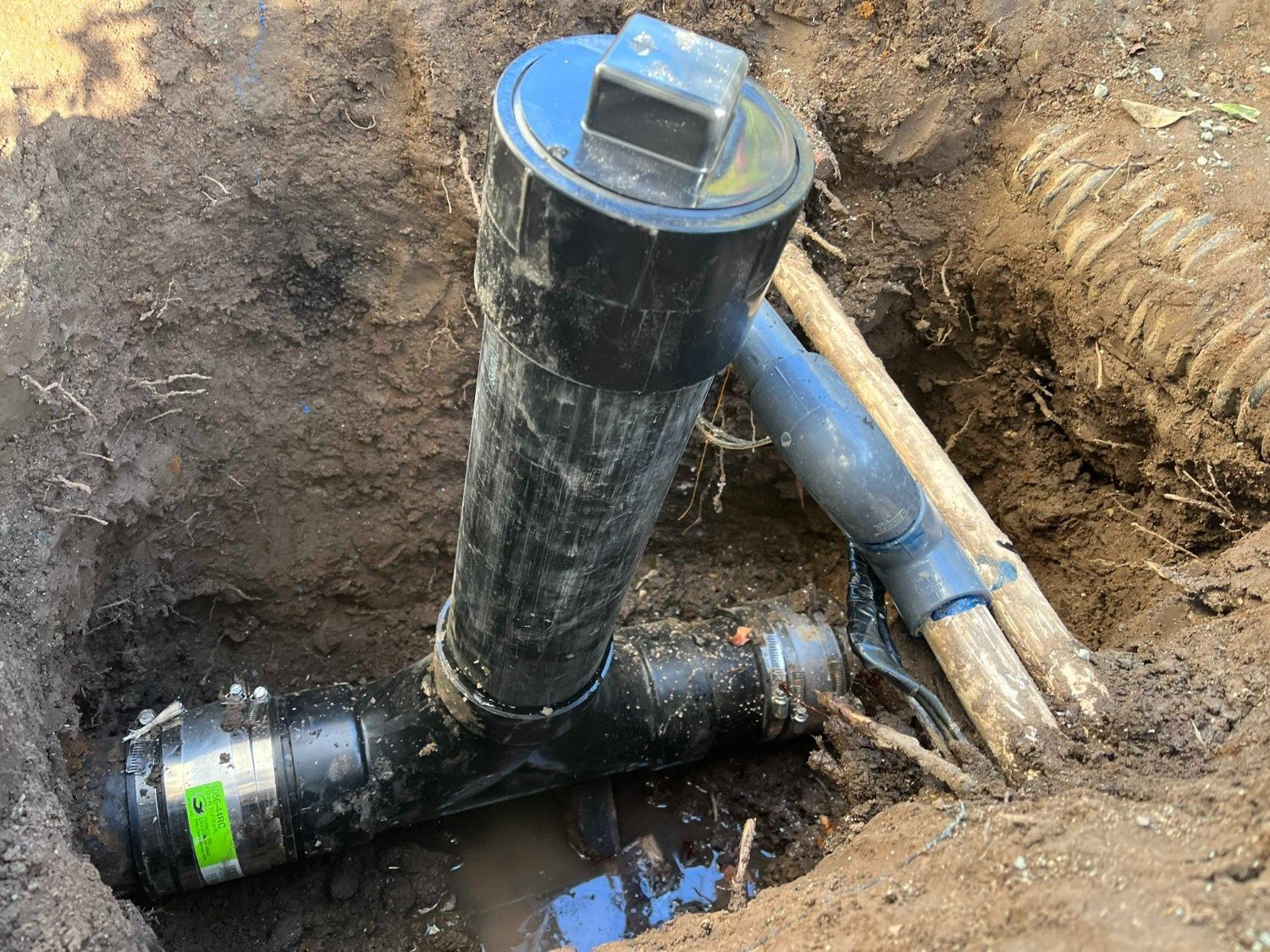 A black plastic cleanout assembly installed in a trench with surrounding dirt and a nearby grey pipe.