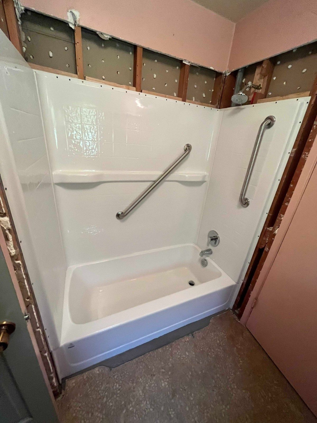 A white bathtub and shower surround with two installed metal grab bars in a bathroom currently undergoing wall repairs.
