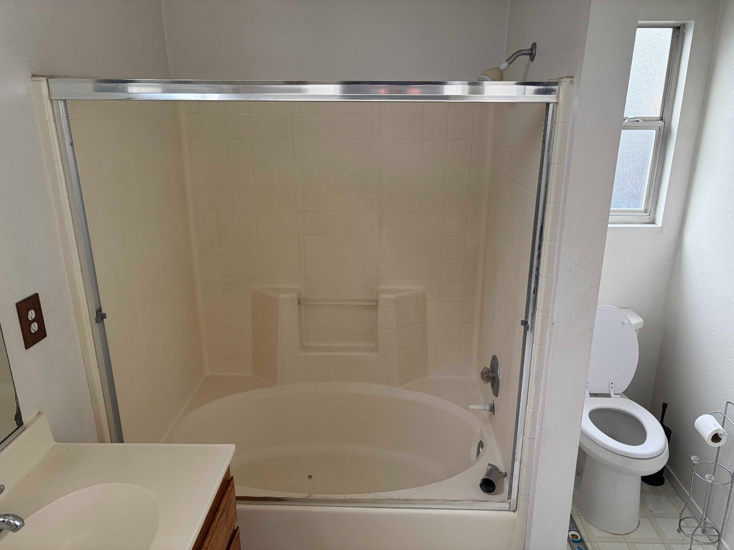 A bathroom with a beige oval tub, shower glass enclosure, a vanity sink in the foreground, and a toilet near a window.