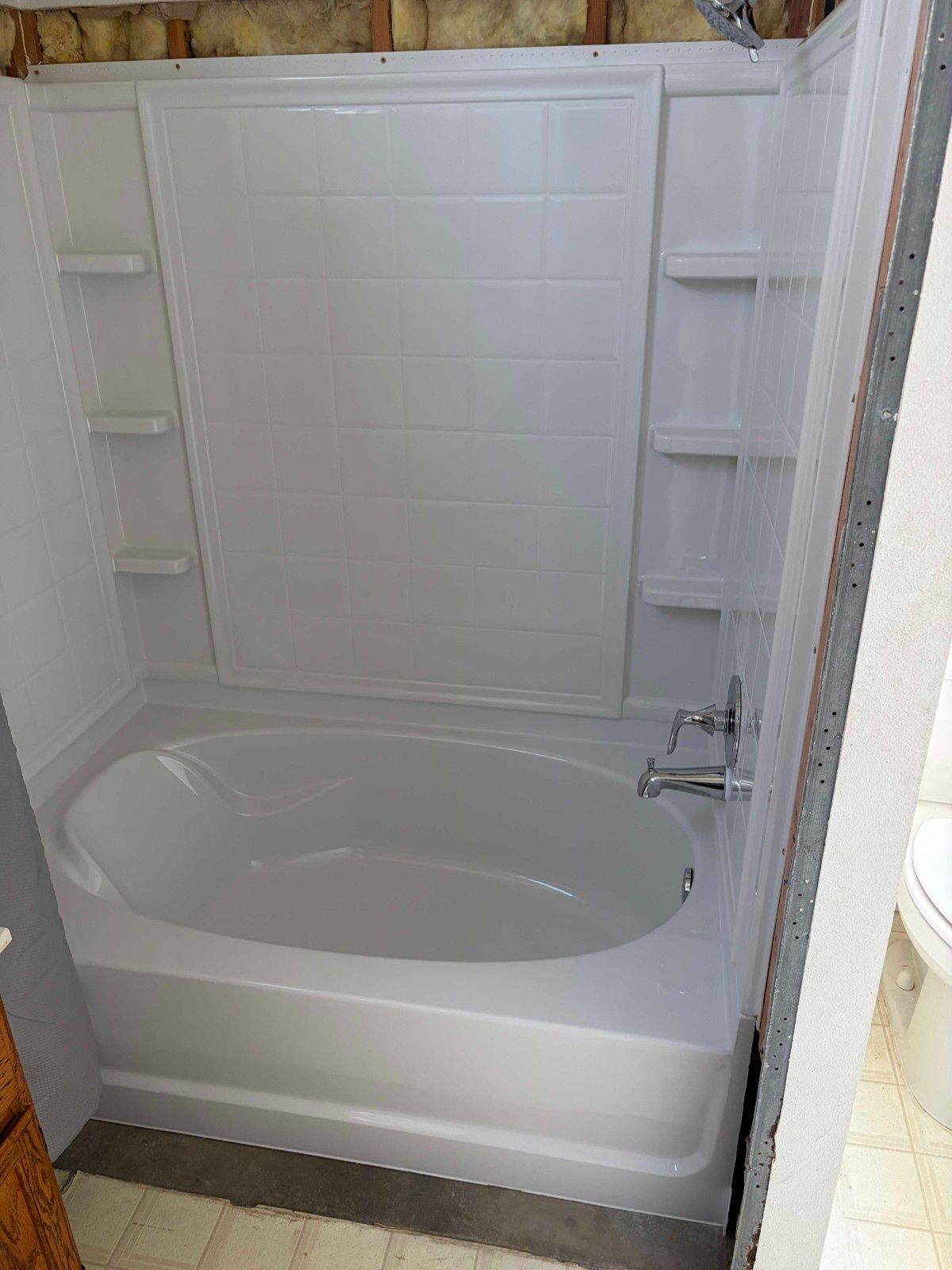A white bathtub and shower surround with built-in shelves in an unfinished bathroom with visible insulation.