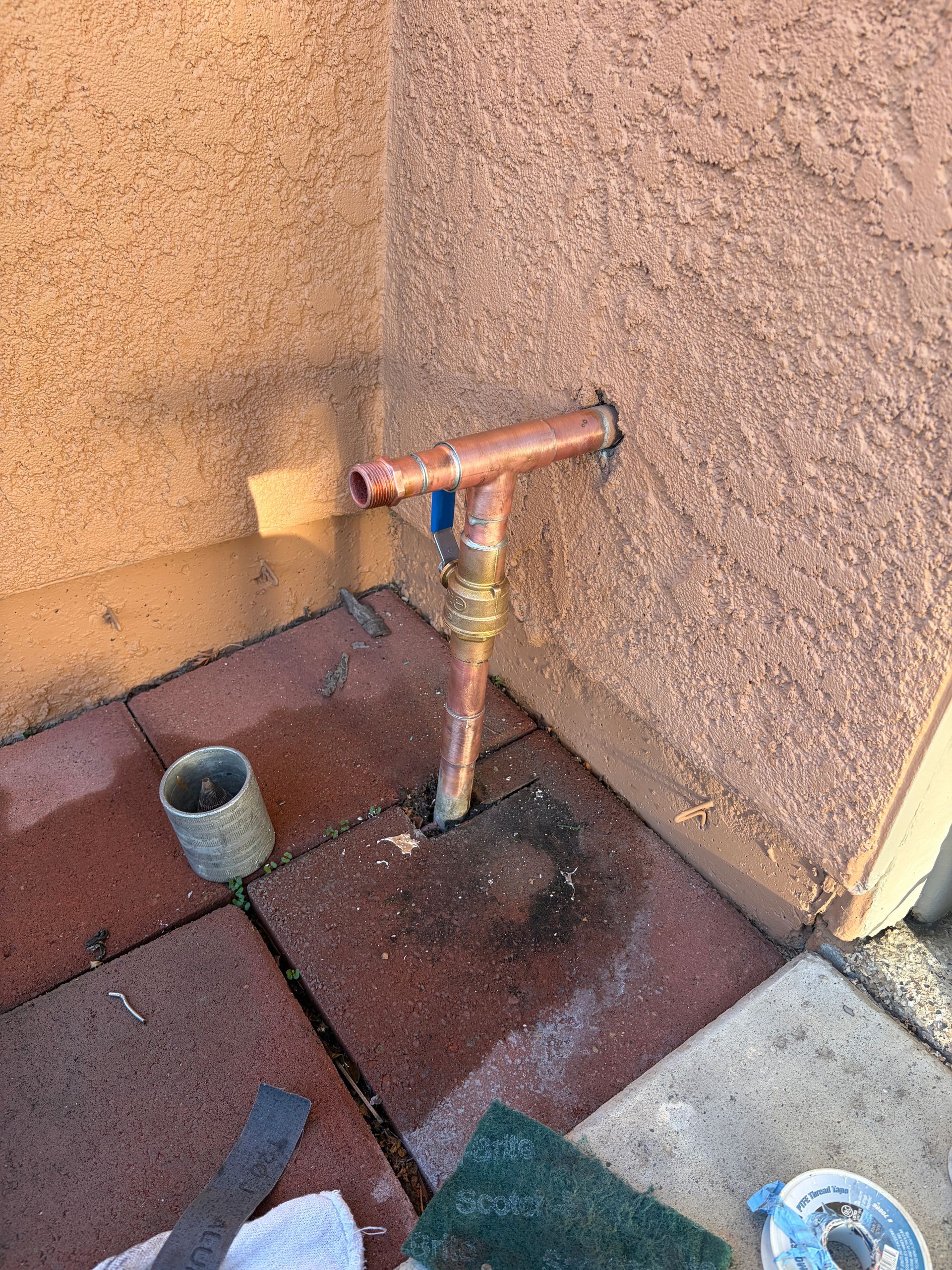 A copper pipe assembly with a ball valve protruding from an exterior stucco wall above a brick patio.