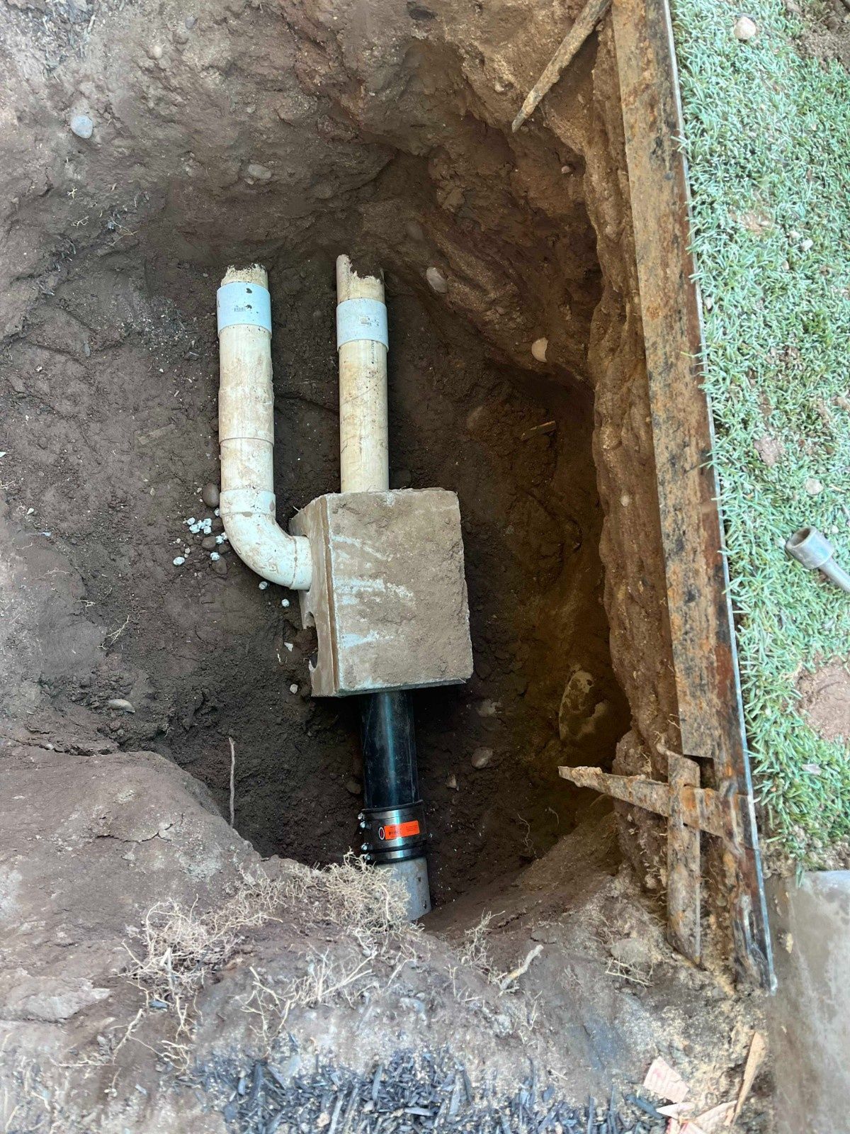 An excavated hole in the ground showing a concrete junction box connected to white PVC and black pipes.