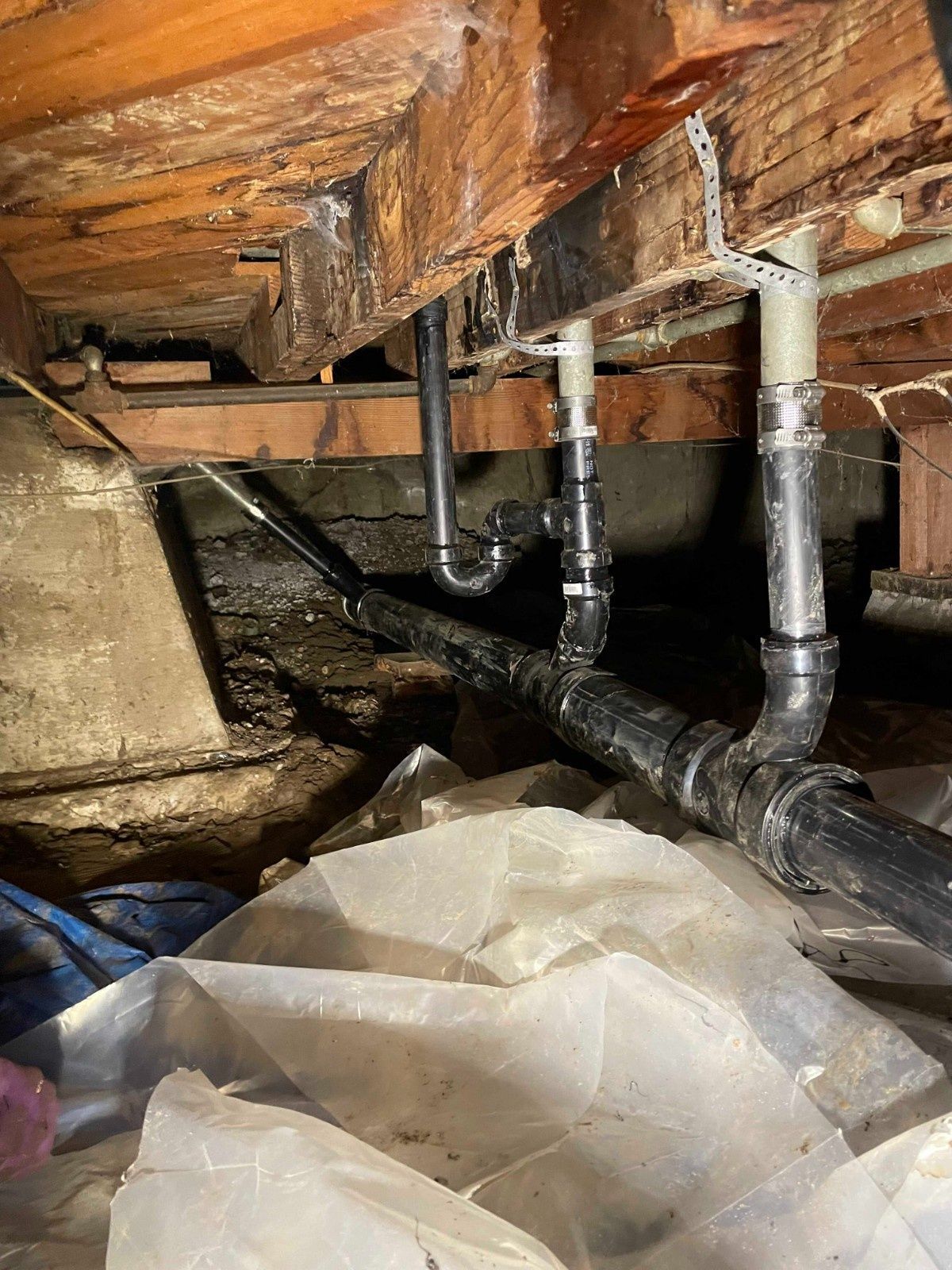 Black plastic plumbing pipes installed underneath a wooden subfloor in a crawlspace.