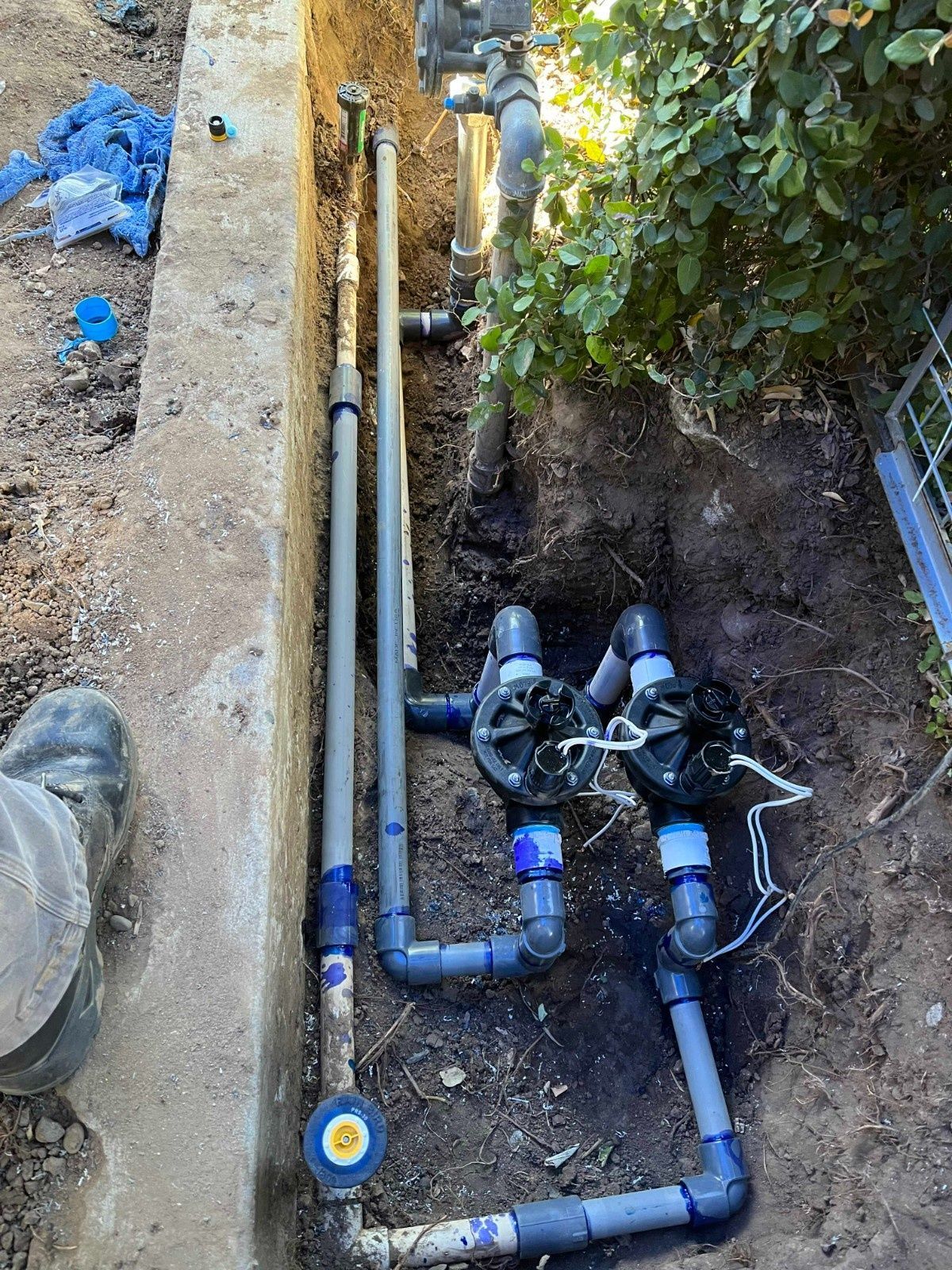 Two irrigation valves and PVC pipes installed in a trench next to a concrete edge.