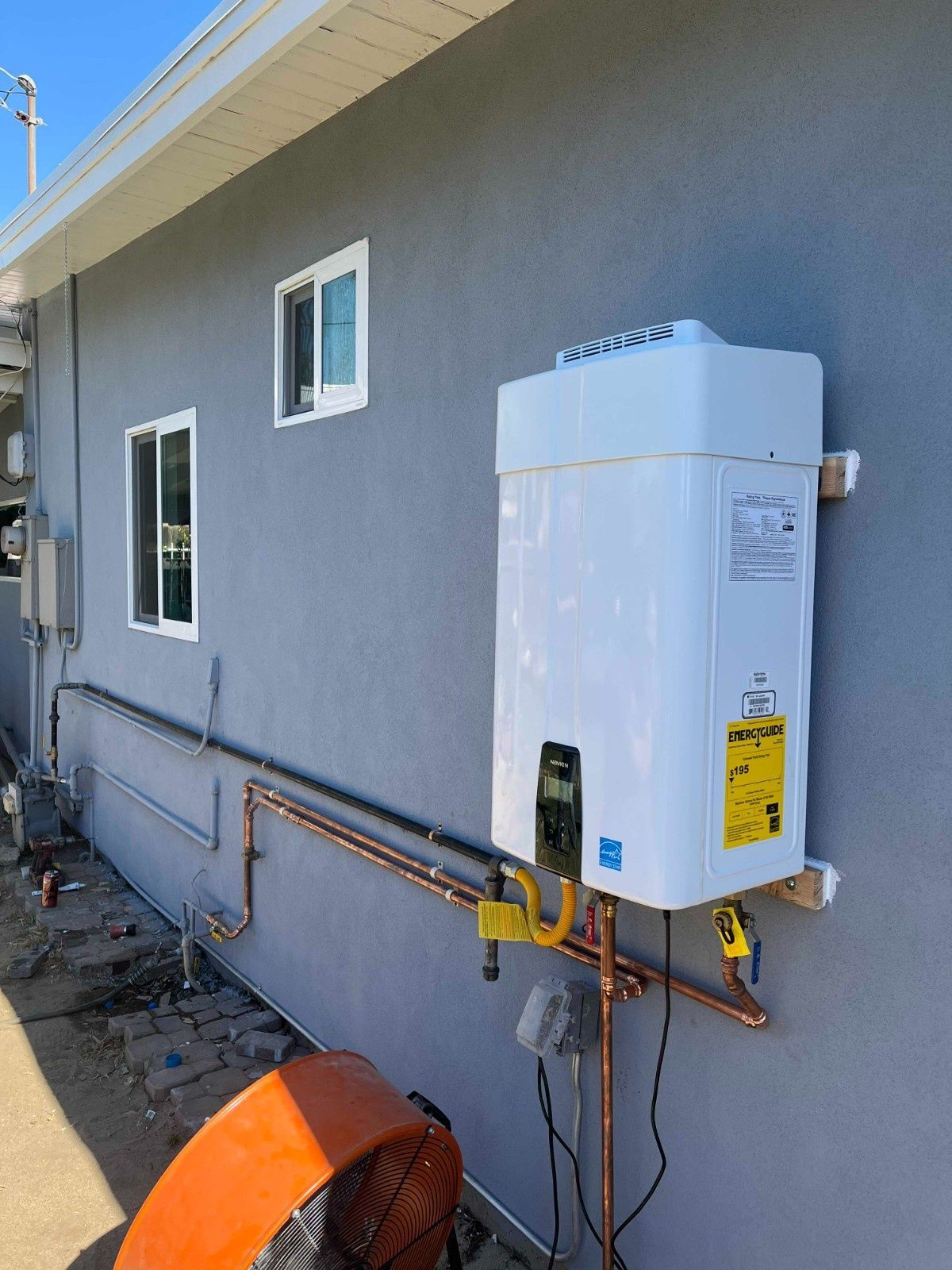 A white tankless water heater mounted on the exterior gray stucco wall of a house with visible copper piping and gas lines.