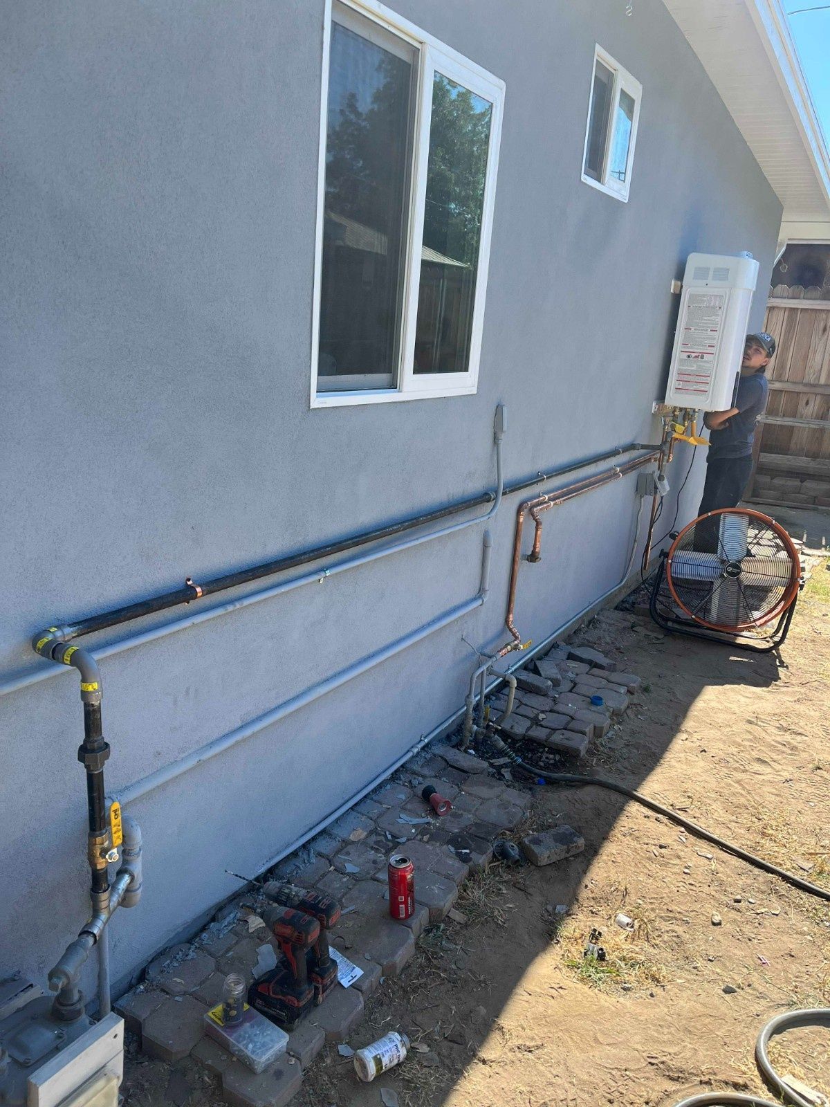 A worker installs a tankless water heater on the exterior wall of a gray house, with gas and water piping running along it.