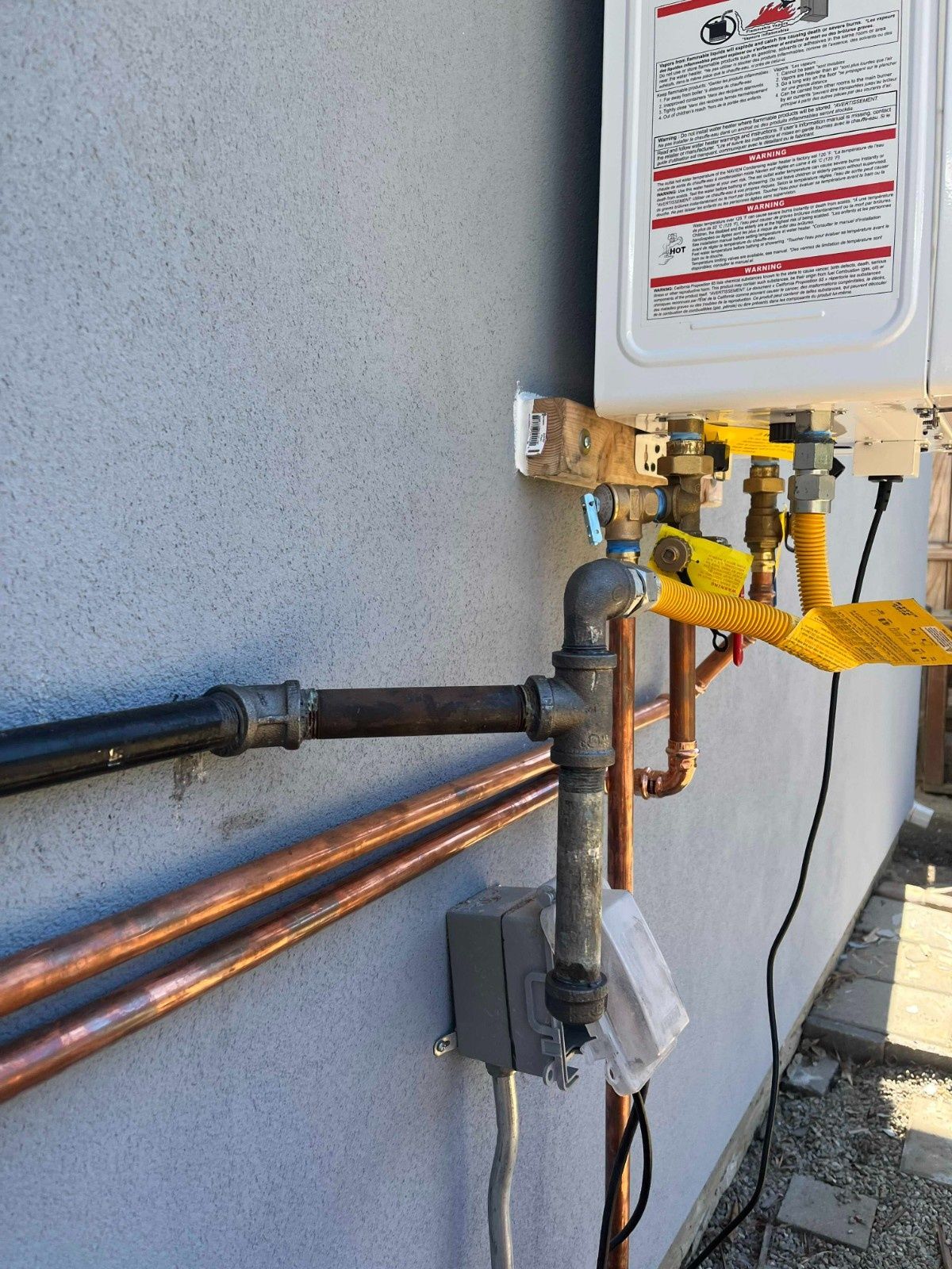 A tankless water heater mounted on a gray wall, connected to black iron gas piping and copper water lines.