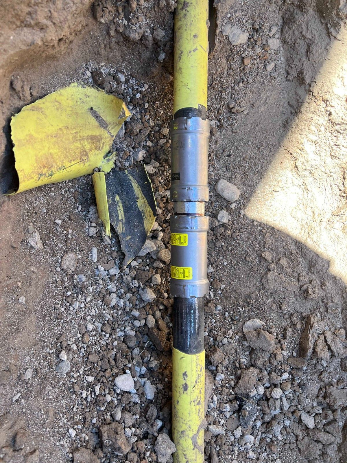 A yellow underground gas pipe with a gray mechanical coupling repair fitting in a dirt trench.