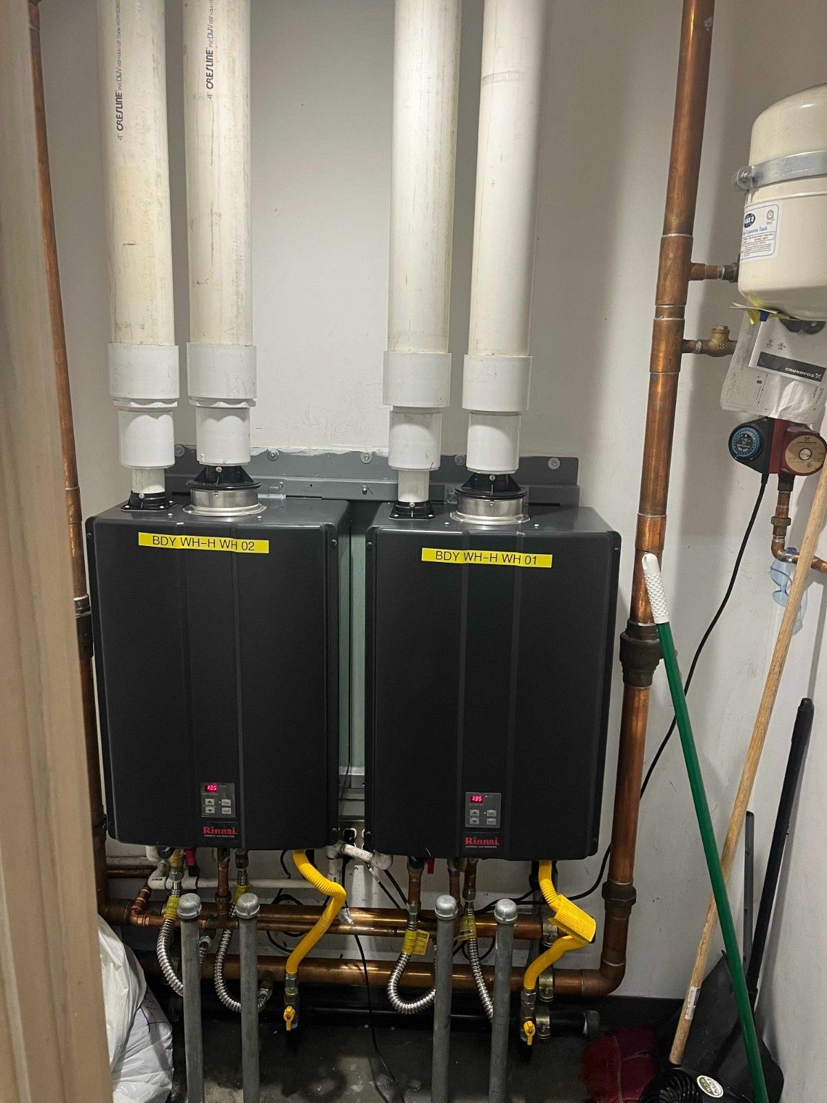 Two black tankless water heaters mounted side-by-side with white vent pipes in a utility closet with copper piping.