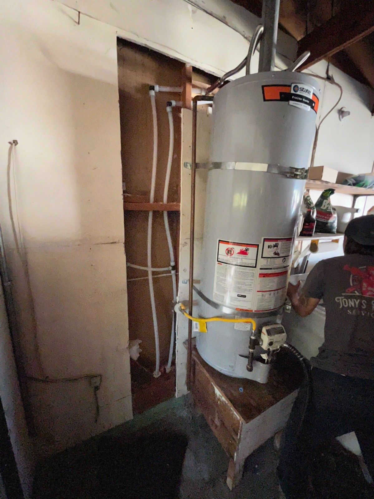 A person installs a gray water heater mounted on a wooden stand in a room with white walls and exposed white plumbing lines.