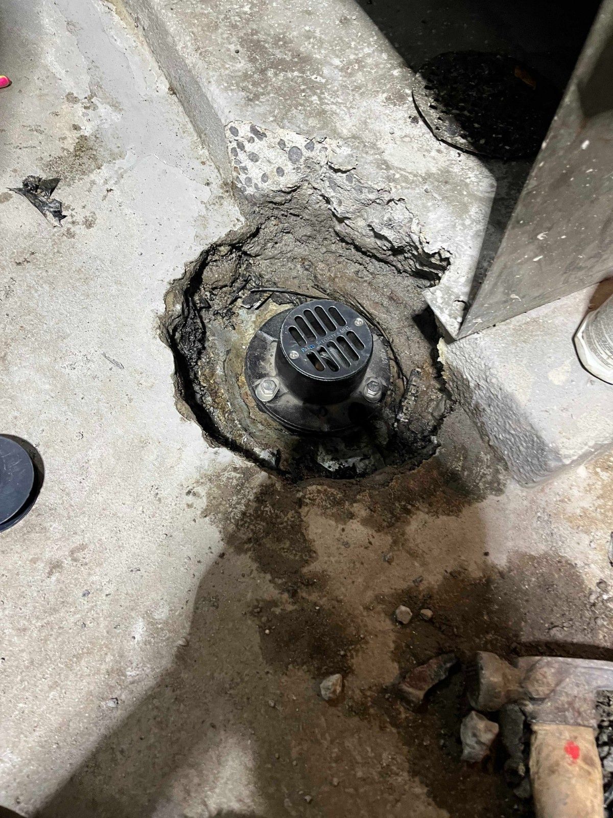 A black circular floor drain is embedded in a partially excavated concrete floor, surrounded by loose dirt and debris.