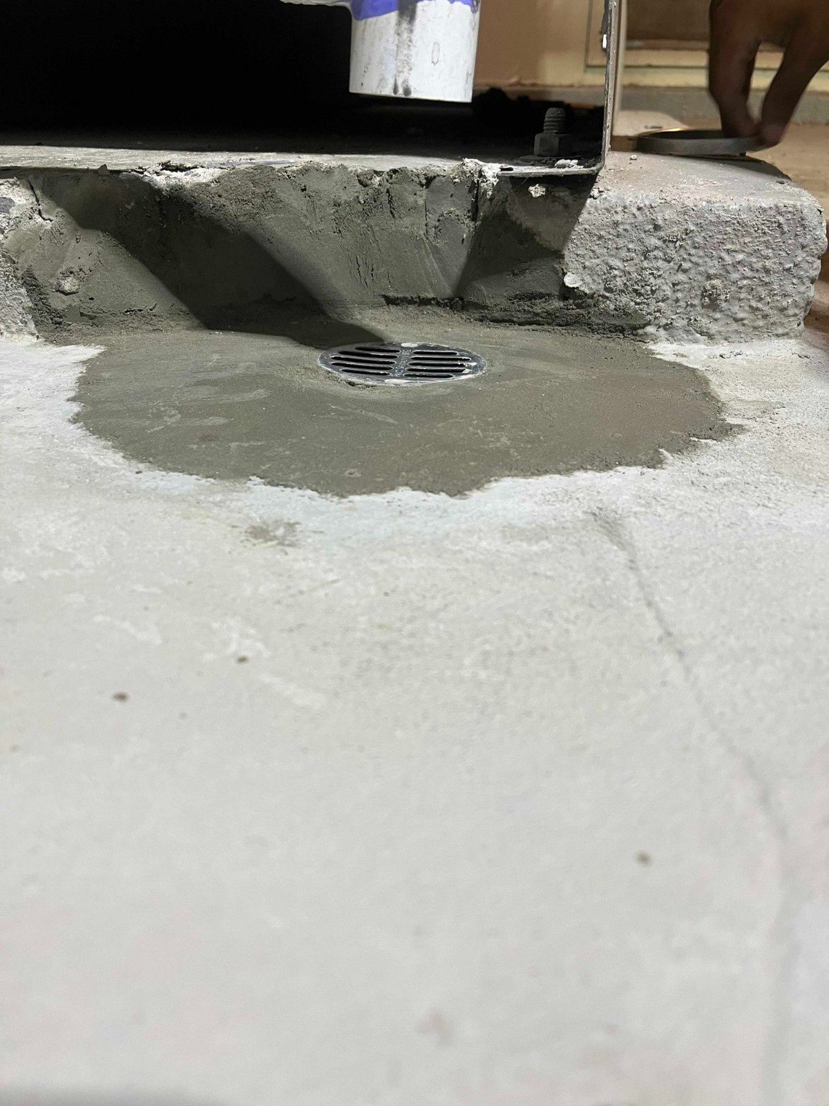 A circular metal floor drain set into a patch of wet concrete near an exterior wall edge.