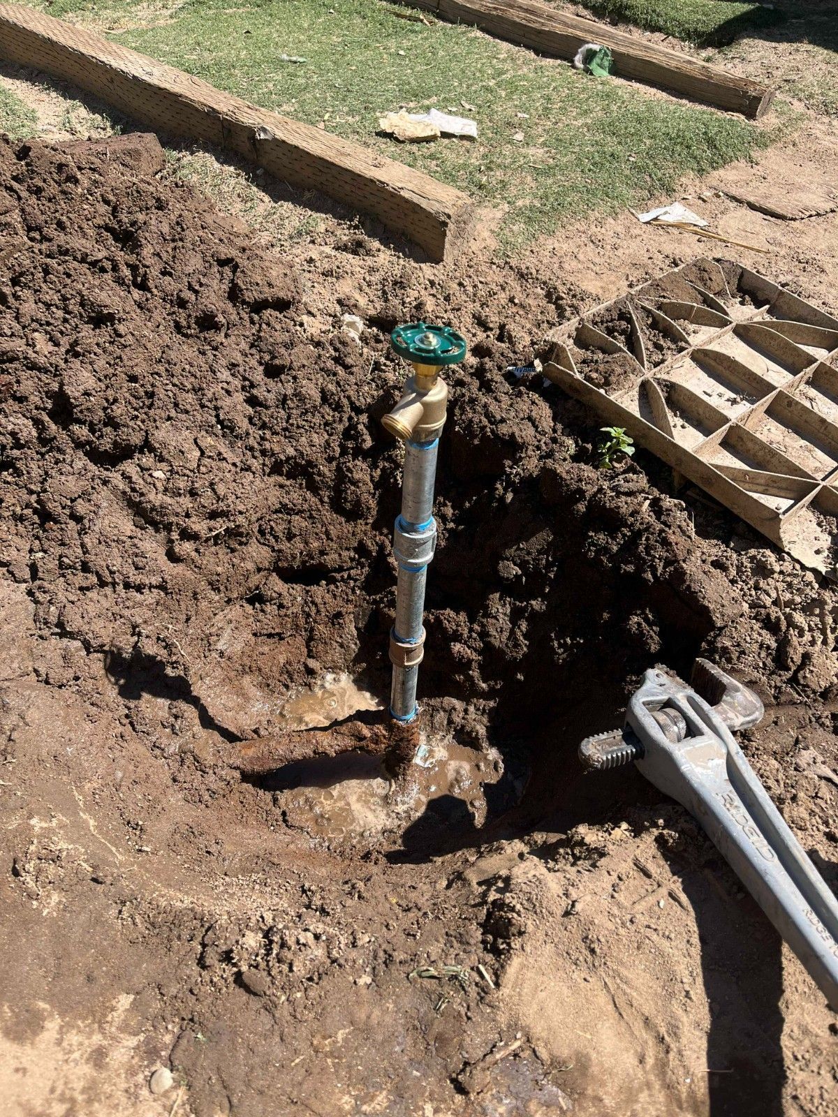 A metal spigot pipe stands upright in a hole dug in dirt, with a large pipe wrench lying nearby on the ground.