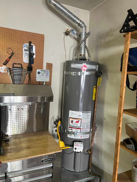 A gray gas water heater stands in a garage next to a metal workbench and wooden shelving unit.