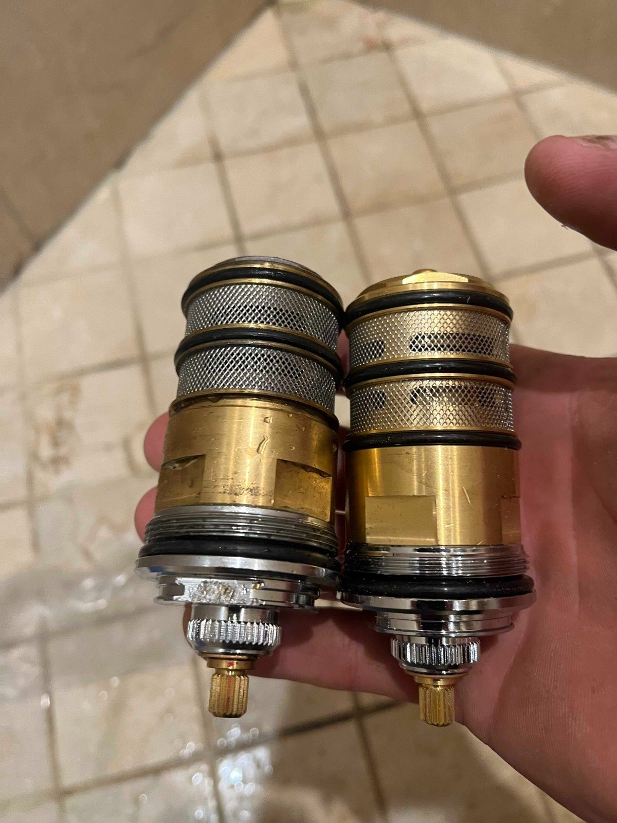 A person's hand holding two brass and silver faucet valve cartridges for comparison.