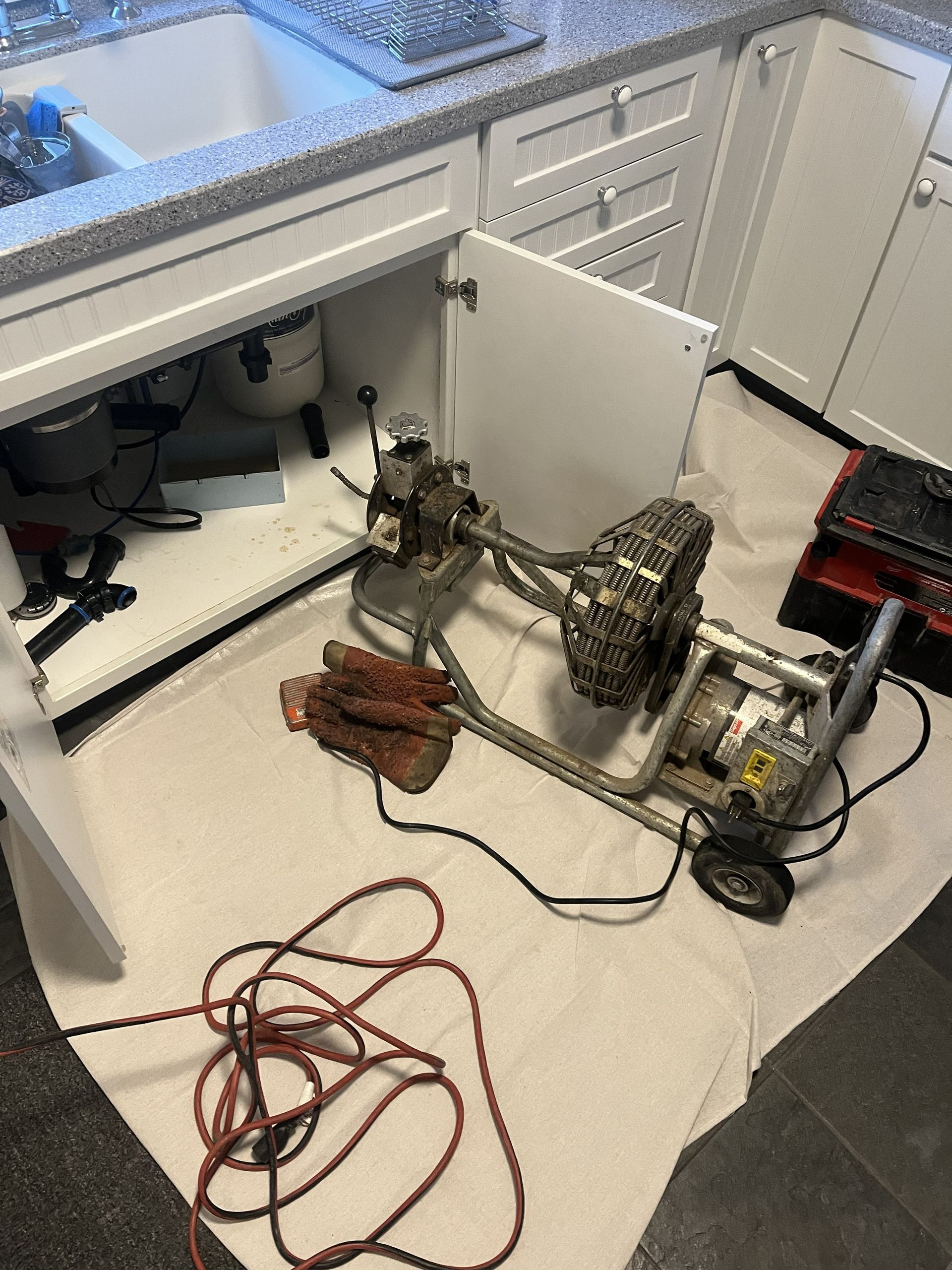 A motorized drain cleaning machine sits on a protective cloth in front of an open kitchen cabinet.