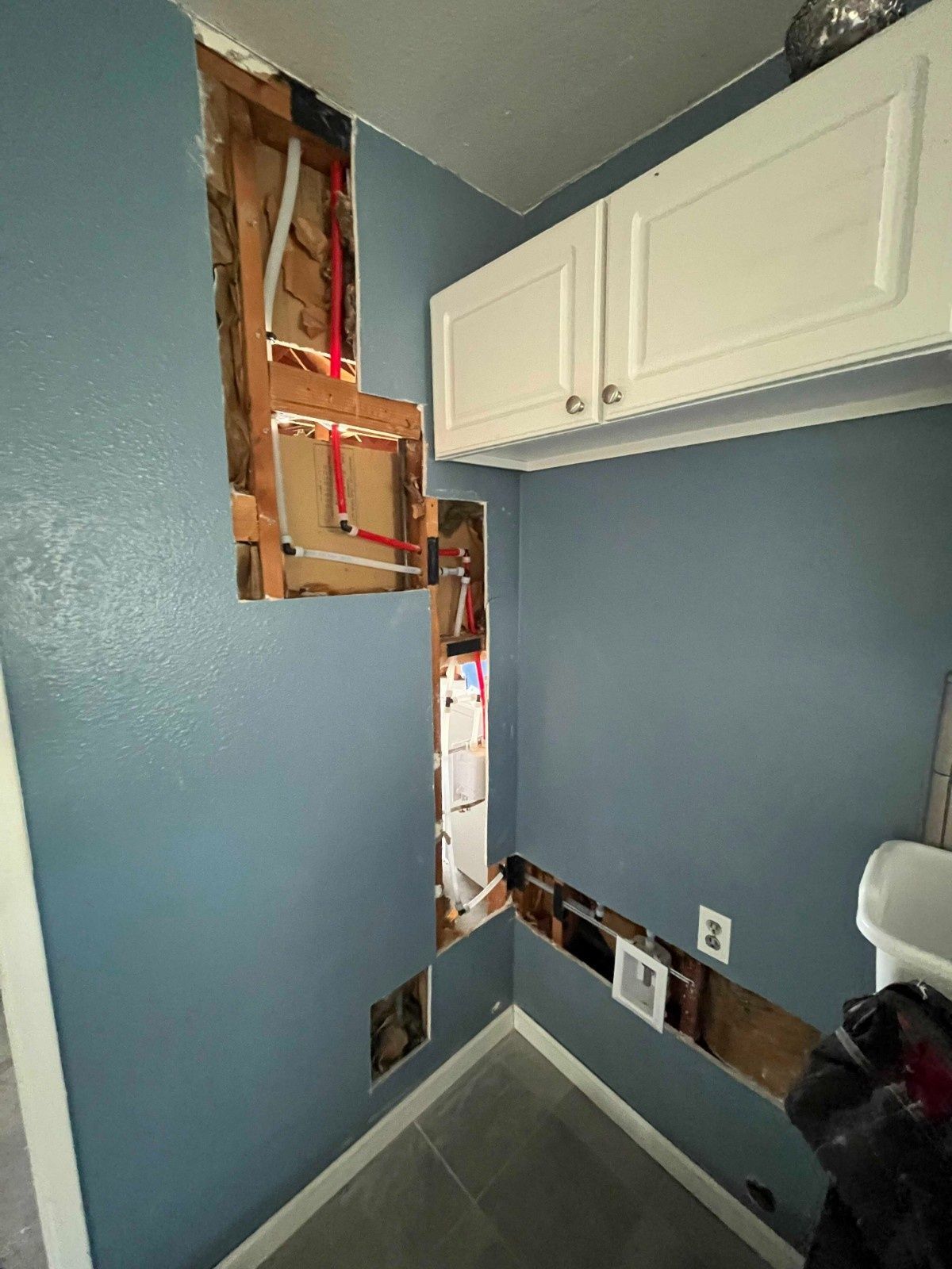 A blue-walled room with large cutouts in the drywall revealing PEX plumbing pipes and electrical wiring inside the studs.