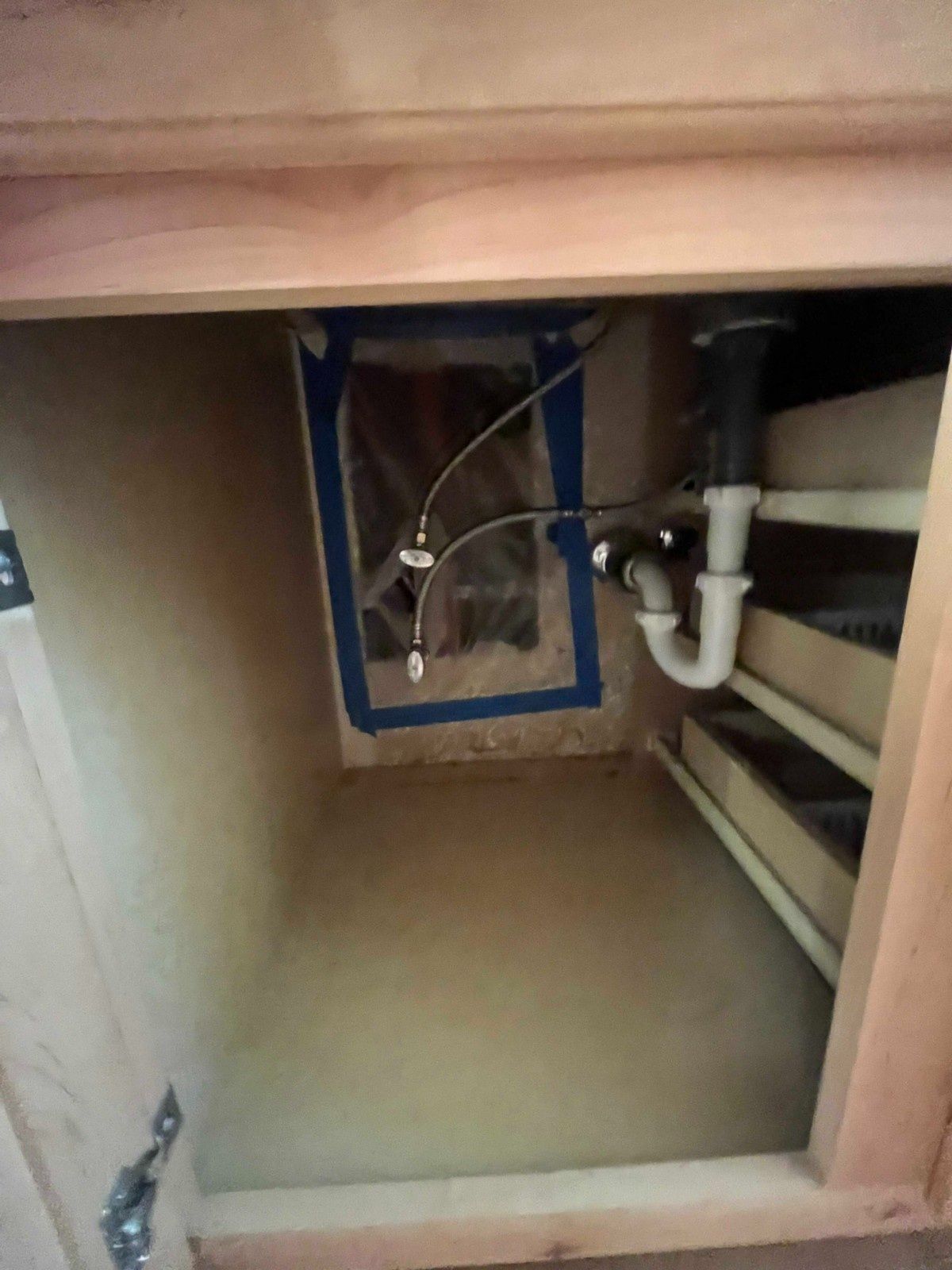 The interior of a wooden cabinet under a sink showing plumbing pipes and supply lines against a wall with blue tape.
