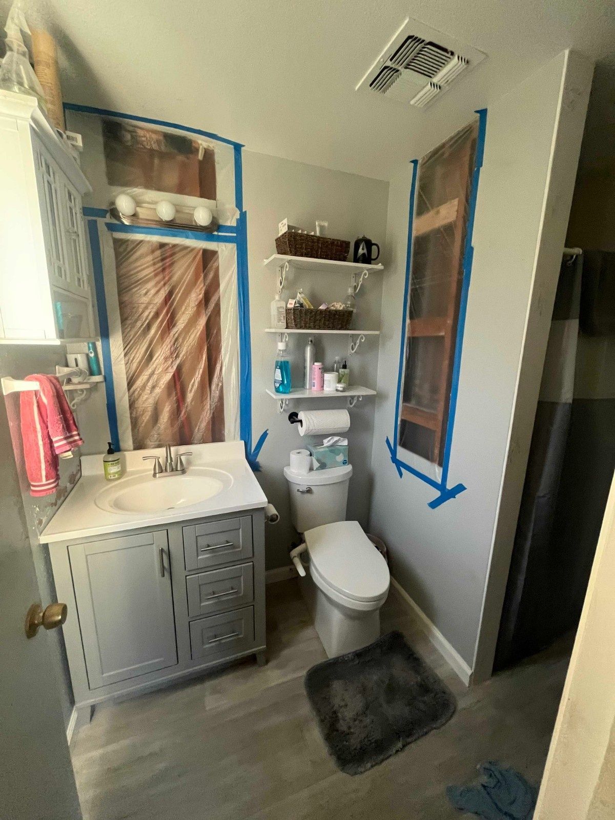 A bathroom with a grey vanity, a toilet, open shelving, and blue painter's tape outlining unfinished wall projects.