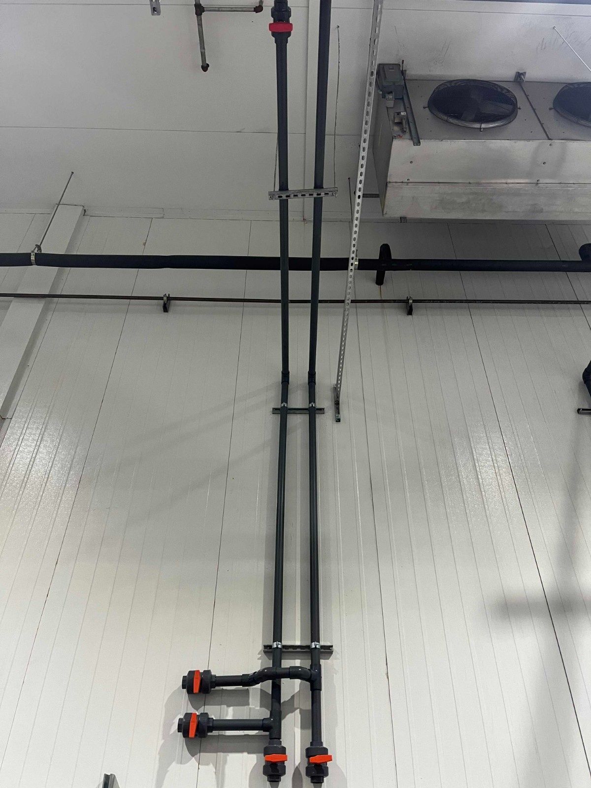Two black pipes run vertically down a white industrial wall, featuring orange-accented valves at the bottom and joints.