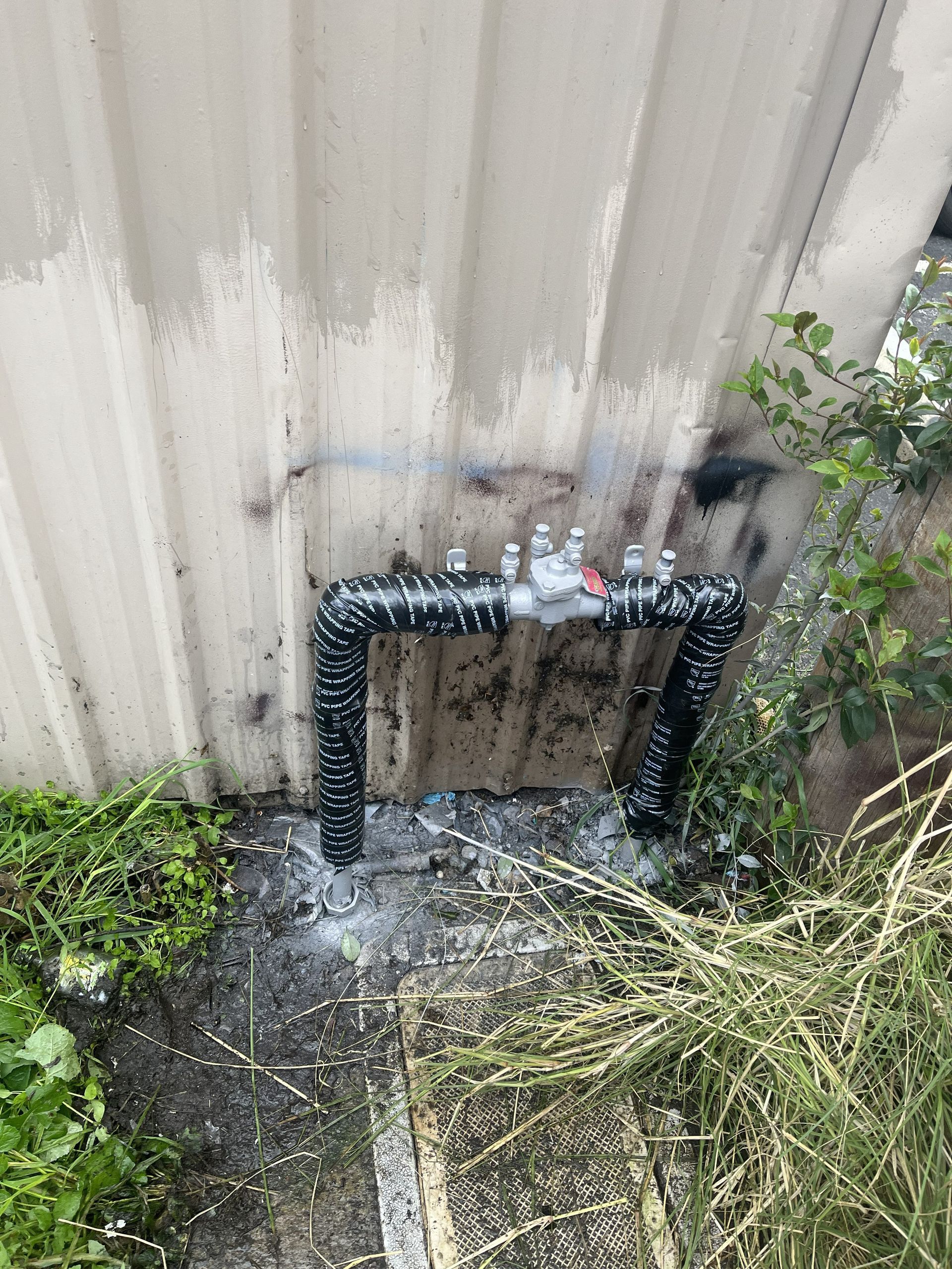 A water pipe assembly wrapped in black protective tape, mounted against a light-colored metal wall exterior.