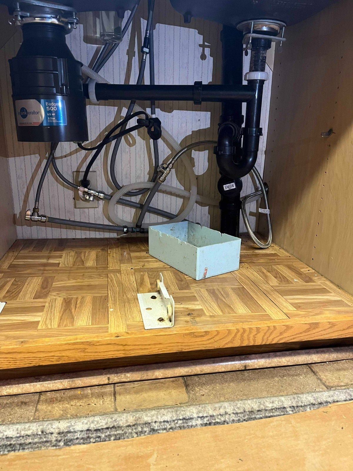 Under-sink view showing a garbage disposal unit, plumbing pipes, and a small light blue bin sitting on a wood floor.