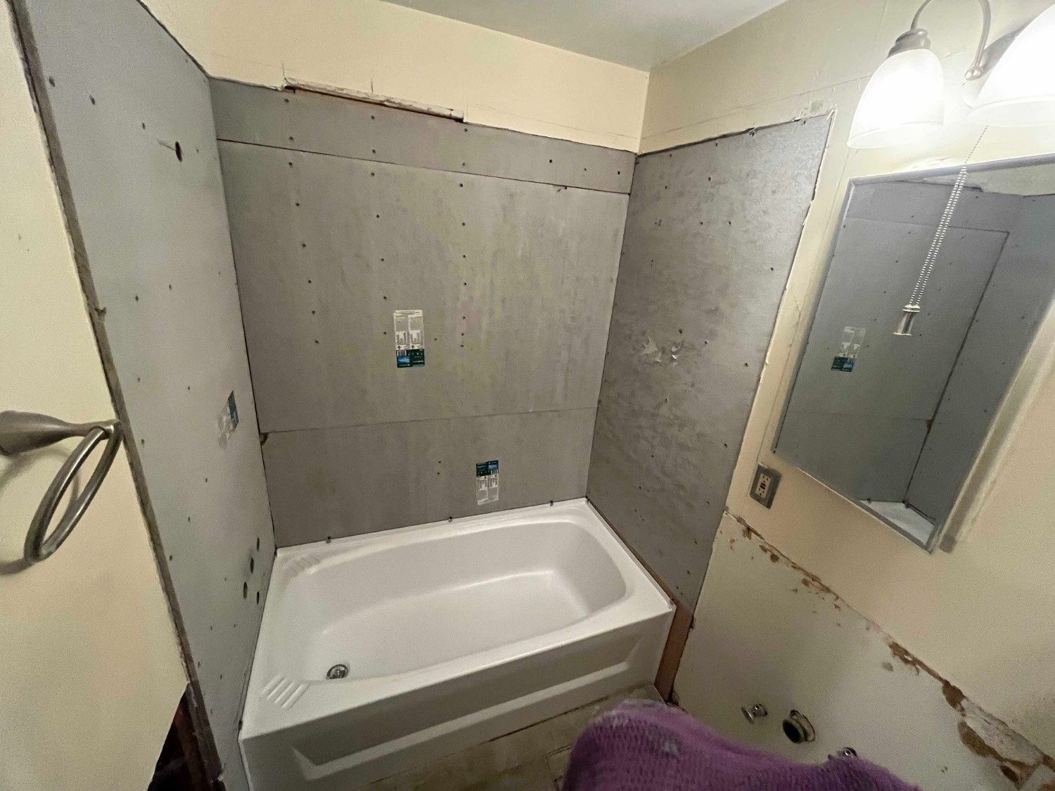 A bathroom undergoing renovation, featuring a new bathtub installed against gray wall panels and an adjacent mirror.